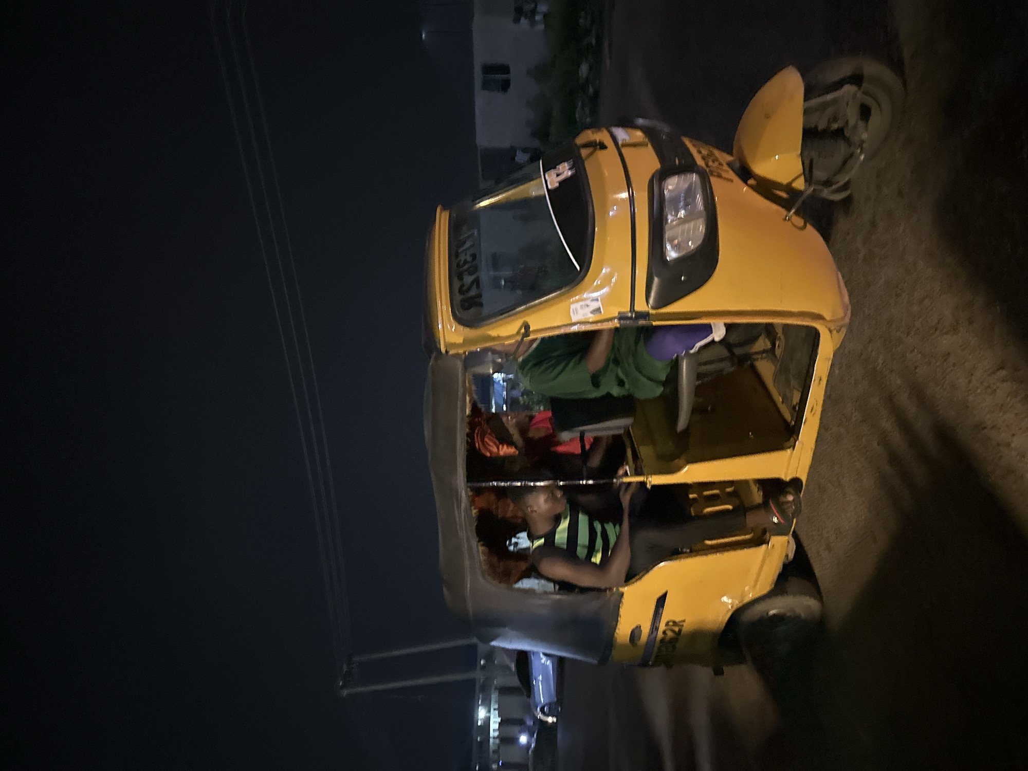 The image shows a yellow auto-rickshaw, commonly known as a "Keke Napep" or "Marwa" in Nigeria, parked on a street at night. Three people are visible inside the auto-rickshaw. The driver, a young man wearing a black and green striped vest, is seated in the front. Behind him, two other individuals are seated, one wearing a green top and another in a red garment. The auto-rickshaw is the primary object, with its yellow body and dark canopy. The scene is dark, indicating nighttime, and there are no visible weather cues. The background shows the faint outline of buildings and utility poles, suggesting an urban or semi-urban setting in Jalingo, Nigeria. The overall mood appears neutral, with no strong emotional expressions evident on the faces of the individuals. Text is visible on the windshield of the auto-rickshaw, appearing as reversed characters, possibly a license plate or registration number. Another piece of text is visible on the side of the auto-rickshaw, reading "3862R".