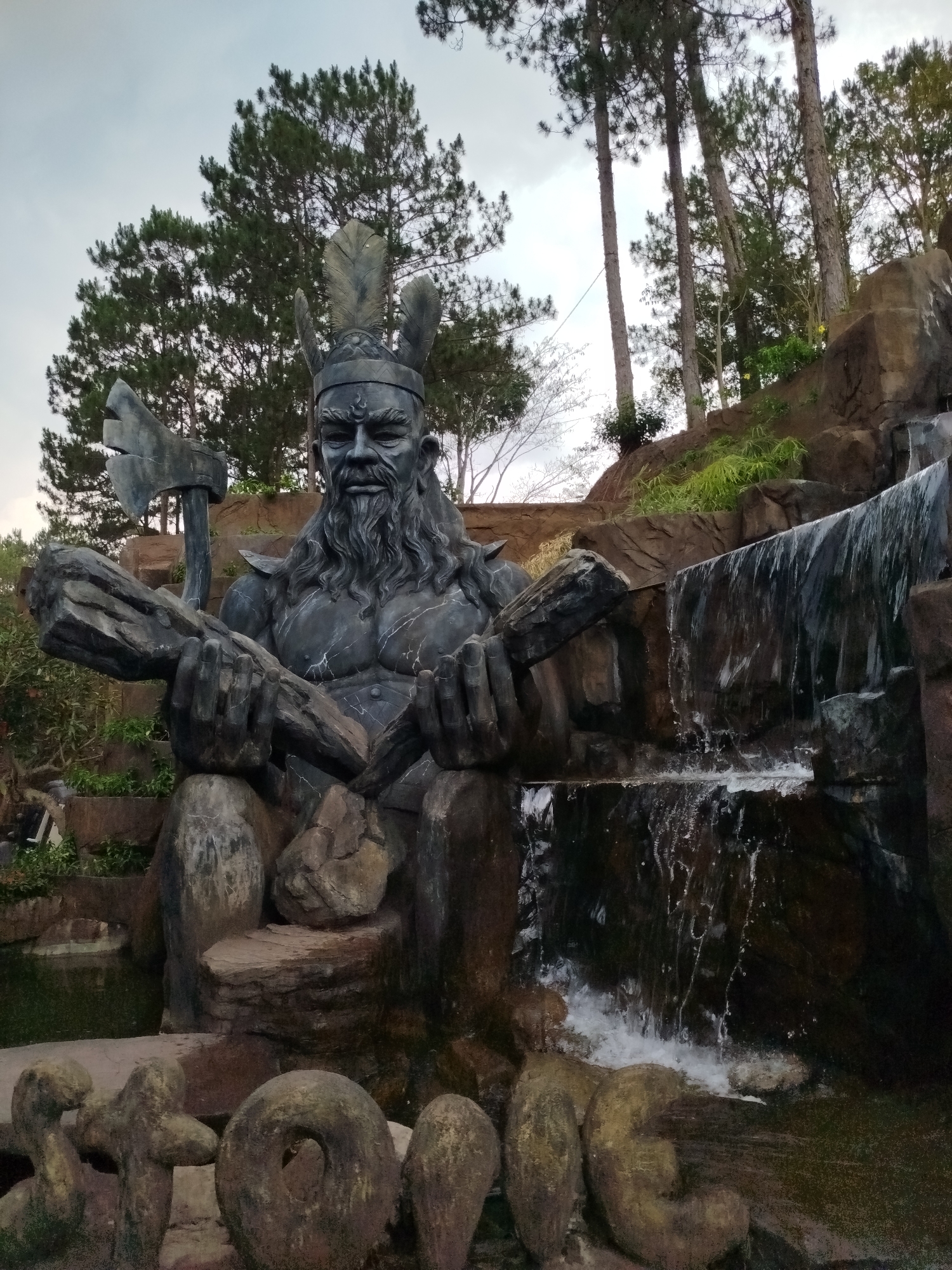 A statue of a bearded man, possibly of Viking or Native American descent, is the focal point of the image. The statue is made of a dark material, perhaps stone or dark-painted concrete, and depicts a muscular man with a stern expression. He wears a headdress with feather-like decorations. In his hands, he holds a rough-hewn log, and an axe rests against his shoulder.
The statue is situated in an outdoor setting, likely a park or garden. Behind the statue is a rocky formation with a waterfall cascading down. The waterfall is small, but the water is clearly flowing. Vegetation, including trees and green plants, surrounds the statue and the rocky formation. The sky is overcast.
The statue is located in the city of Da Lat, Vietnam.