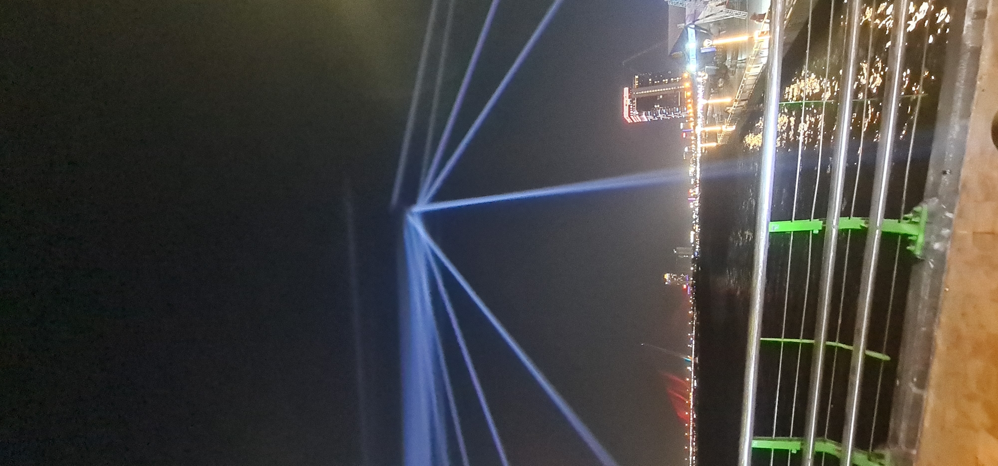 This nighttime image captures a vibrant light show illuminating the sky over a body of water in Da Nang, Vietnam. Numerous intense beams of light, predominantly blue and white, crisscross the dark sky, originating from the far bank of the river. These lights create striking linear patterns, with one prominent horizontal beam stretching across the mid-ground and other diagonal rays converging at a central point.
Across the water, a city skyline is visible, bustling with various illuminated buildings and structures. The lights from the city and the light show reflect on the water's surface, adding to the spectacle. In the immediate foreground, a metal railing or barrier is prominent, consisting of multiple vertical bars interspersed with horizontal, bright green supports. A wooden surface is visible at the bottom right, suggesting the perspective is from a bridge or an elevated viewing platform. The scene depicts a clear night with no visible weather obstructions, highlighting a public event or a nightly urban light display. There is no visible text in the image.