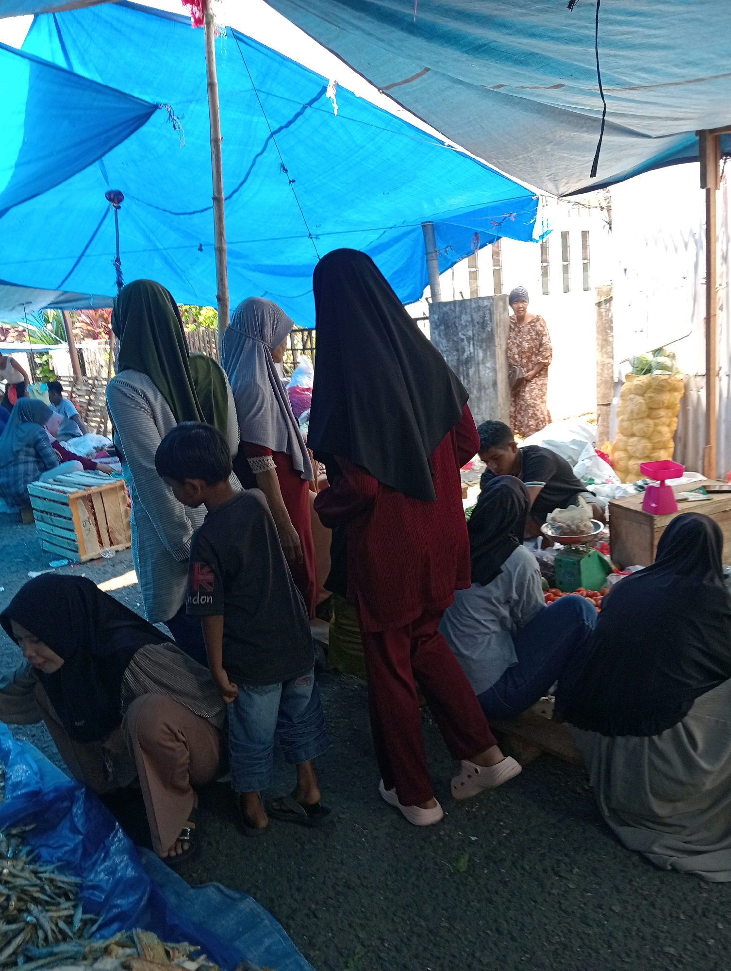 The image depicts an outdoor market scene in Pariaman, Indonesia, likely a roadside or open-air market. A large blue tarp, supported by wooden poles, provides shade over the area. Several people, mostly women wearing hijabs, are present, some engaged in selling or buying goods. In the foreground, a young boy in a dark t-shirt stands, while a woman in a black hijab crouches near a blue tarp filled with dried fish. Further back, a man is attending to produce, with tomatoes and other vegetables visible. To the right, stacks of yellow produce, possibly onions or similar items, are piled high. The ground appears to be paved or gravel. A woman in a floral dress stands in the background, near a building. The overall atmosphere suggests a typical market day.