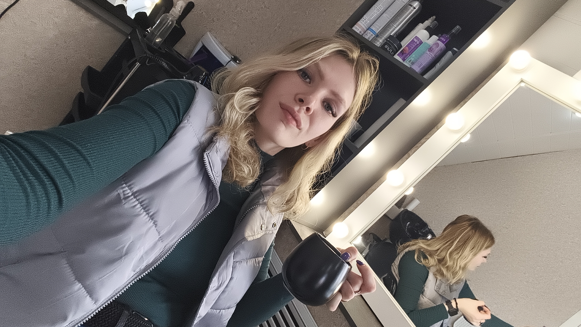 A young blonde woman is captured in a close-up selfie, looking directly at the camera with a confident expression. Her wavy, shoulder-length hair frames her face. She wears a dark green long-sleeved top underneath a light grey quilted or puffer vest. In her right hand, she holds a dark, possibly black, ceramic mug, with her fingernails painted a deep purple hue.
The setting appears to be an indoor space, likely a salon or a dressing room, based on the background elements. To her right, a large vanity mirror with exposed round light bulbs along its frame reflects her image from a different angle, showing her in profile, looking downwards and engaged in an unseen activity. Behind her, shelves are visible, filled with various hair care products and styling tools, indicating a professional environment. Text on some product bottles, like "shuurr" and "expo," is partially discernible. The walls are a textured light beige or off-white. The overall lighting is bright and even, typical of indoor artificial light. The image provides a glimpse into a moment of casual self-portraiture within what seems to be a workspace in Русский Вожой, Russia.