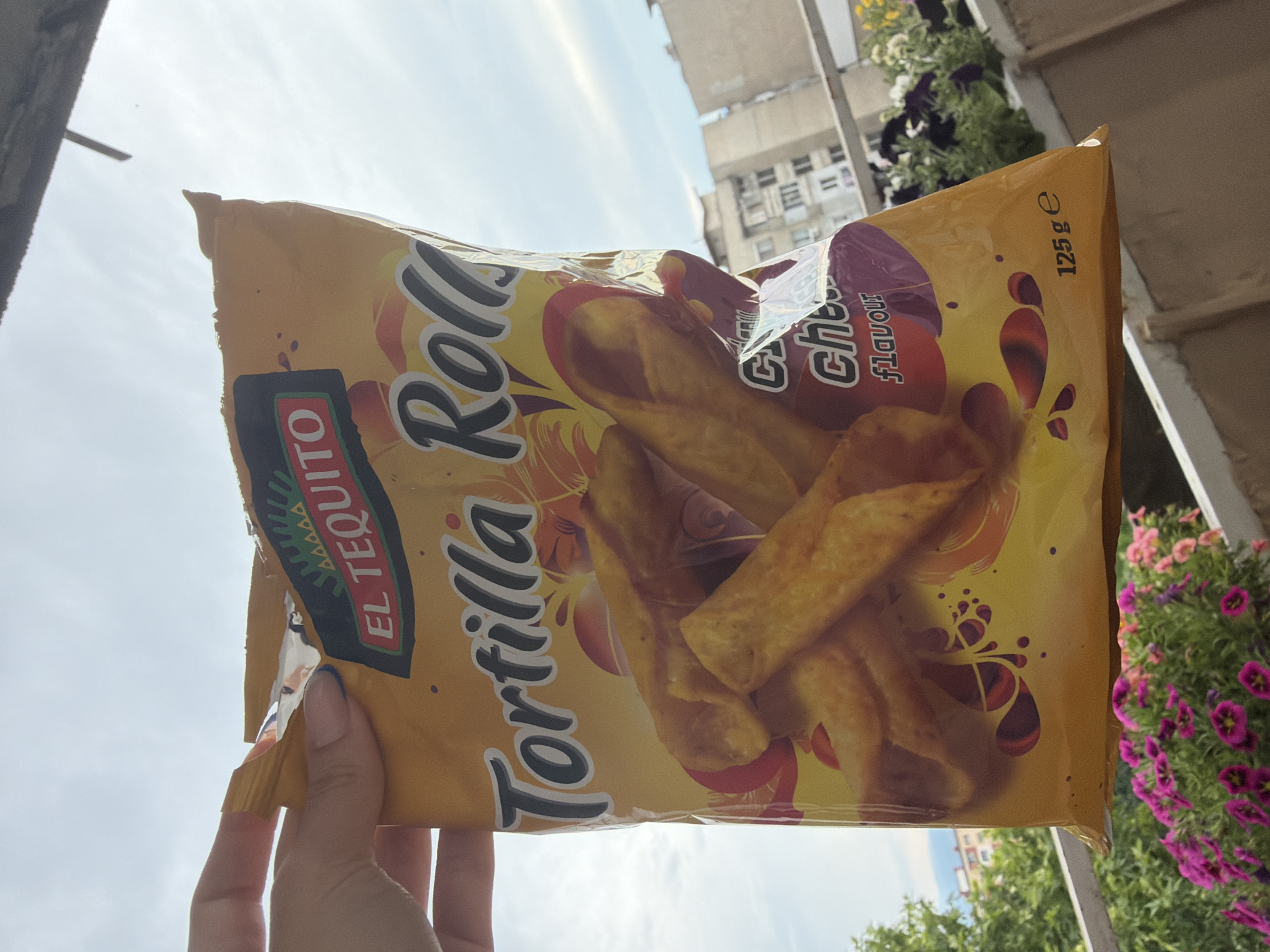 A hand holds a bag of "Tortilla Rolls" snack chips with a "Cheese Flavour" by "EL TEQUITO". The bag is yellow with red and purple accents and images of the rolled chips. The background shows a cloudy sky, apartment buildings typical of Pazardzhik, Bulgaria, and a railing with potted purple and pink flowers. A portion of the top of the bag appears torn open. The bag indicates a net weight of 125g.