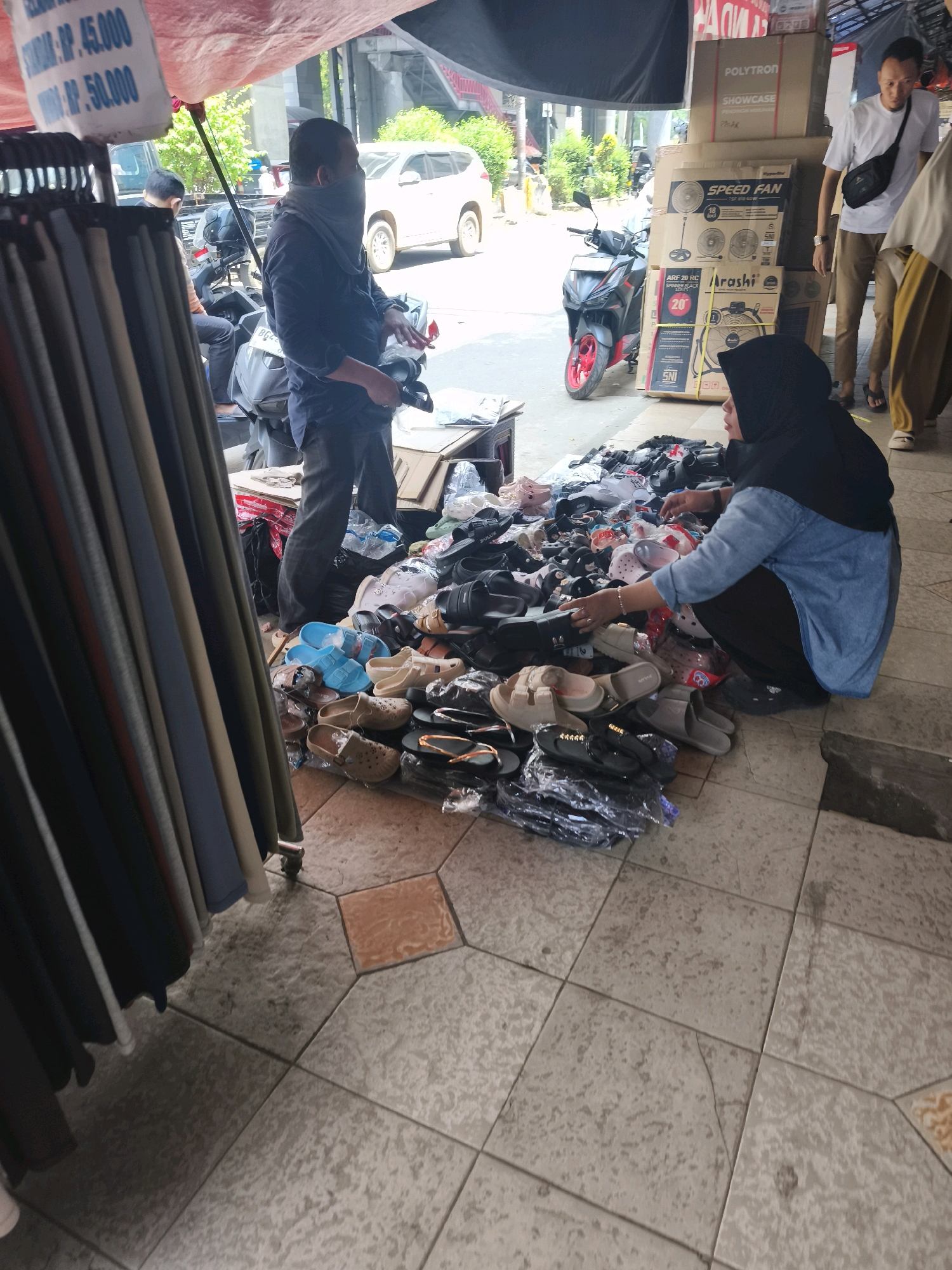 The image captures a street-side market scene in Kelurahan 16 Ilir, Kec. Ilir Timur I, Indonesia. In the foreground, a rack of clothing hangs on the left, with various colored garments displayed. To the right, a woman wearing a black hijab and a blue denim shirt is crouched down, sorting through a large pile of footwear, primarily sandals and flip-flops, spread across the tiled ground. Behind her, several boxes are stacked, some labeled with brand names like "POLYTRON" and "SPEED FAN."
A man wearing a dark shirt and a grey face covering stands near a motorcycle, holding what appears to be a small item in his hand. Another man in a white t-shirt and brown pants is visible further back, looking towards the boxes. A white SUV and a black motorcycle with red accents are also present in the background, suggesting a busy urban environment. The overall setting is outdoors, with a makeshift roof or awning providing shade over the market stall.