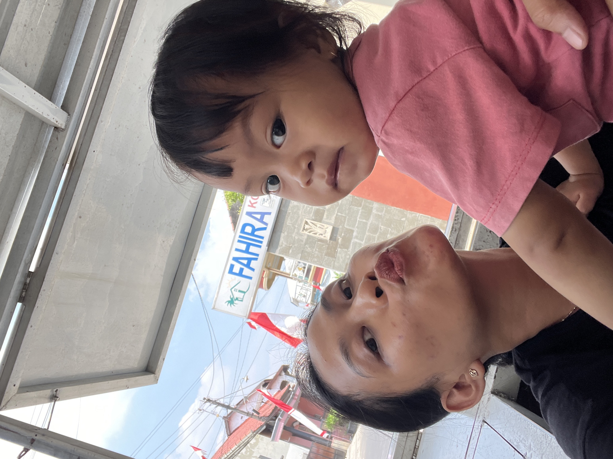 A woman is taking a selfie outdoors in front of a building with a sign that says "FAHIRA" in Kecamatan Pati, Indonesia. She is holding a baby, who is looking at the camera. The woman is making a kissy face, and the baby has a neutral expression. The background shows a street with houses and power lines. The sky is blue and there are some clouds. The overall scene is casual and informal.