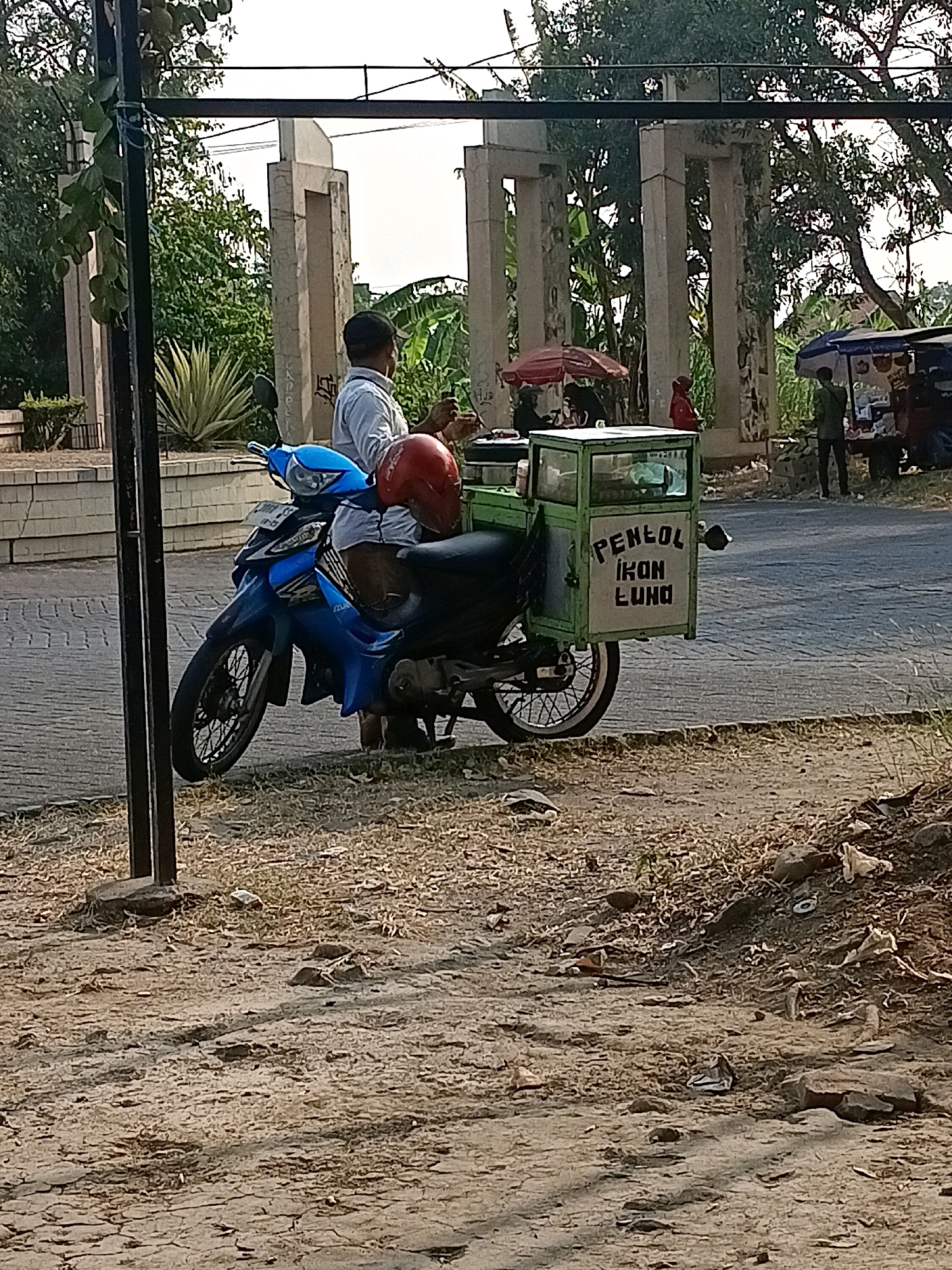 The image shows a man sitting on a blue motorcycle with a green food cart attached to the back. The food cart has text on it that reads "PENLOI IKON LUND". The man is wearing a light-colored shirt. A red helmet is visible on the back of the motorcycle seat. In the background, there are several large stone structures that appear to be part of a monument or historical site. Trees and vegetation surround the structures, and a street is visible with a few people and market stalls. The lighting suggests it is daytime, possibly late afternoon, with a warm, natural light casting shadows. The overall scene depicts a common sight of a food vendor operating from a motorcycle in a semi-urban or rural Indonesian setting, likely in Tawangsari given the location context. The ground in the foreground is dry and dusty, with scattered debris, indicating a somewhat unkempt area.