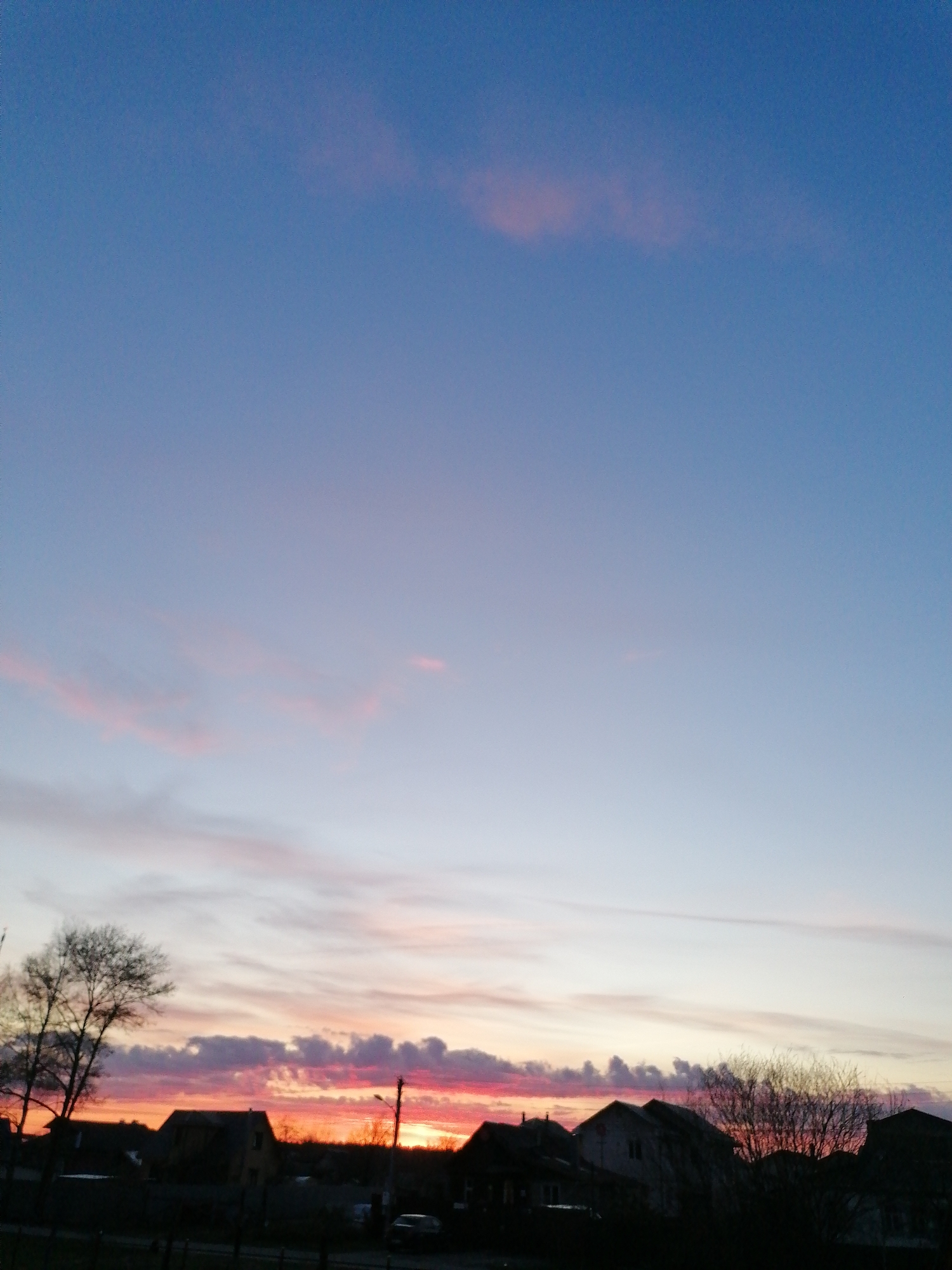 The image depicts a twilight scene with a view of the sky transitioning from blue at the top to shades of pink, orange, and red near the horizon. The lower portion of the image shows a silhouetted cityscape, with trees and buildings visible. A streetlight is positioned to the right. A vehicle is also visible. The location is Unknown, Unknown.