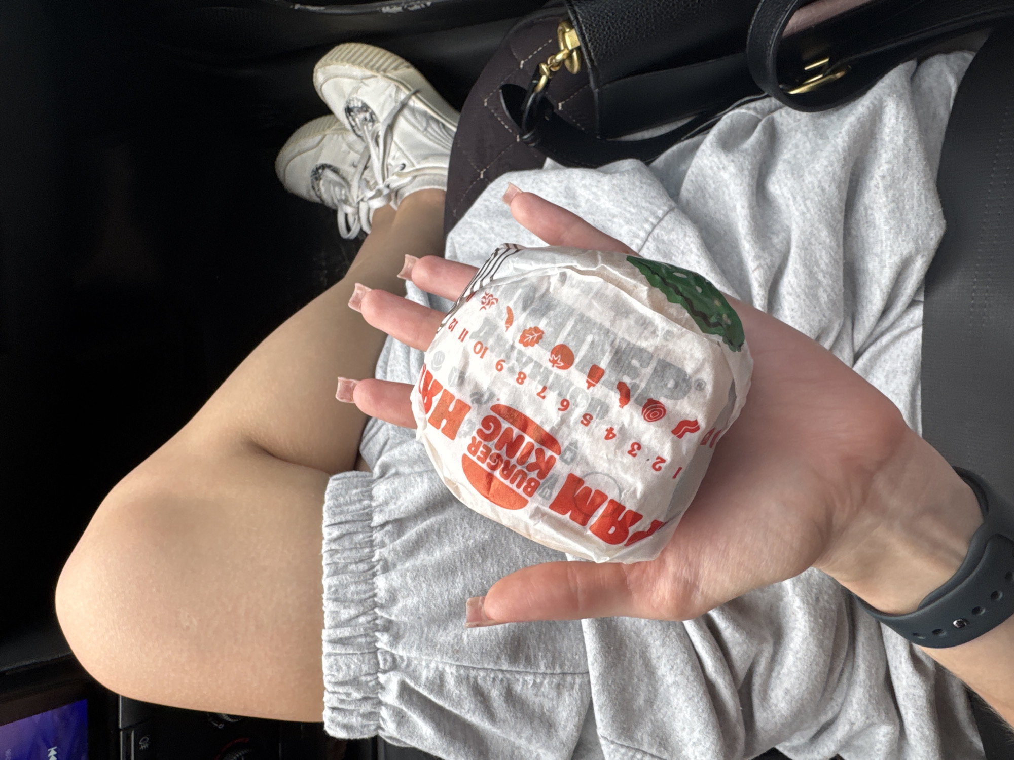 In a vehicle, likely parked, a person is holding a Burger King hamburger wrapped in white paper with red lettering. The person's hand is in the foreground, and they have long, square-shaped fingernails. They are wearing a grey sweat suit and white sneakers. The background shows a dark interior of a car, with part of a black purse visible. There is no information or visual cue to place the scene in Selishcha, Russia.