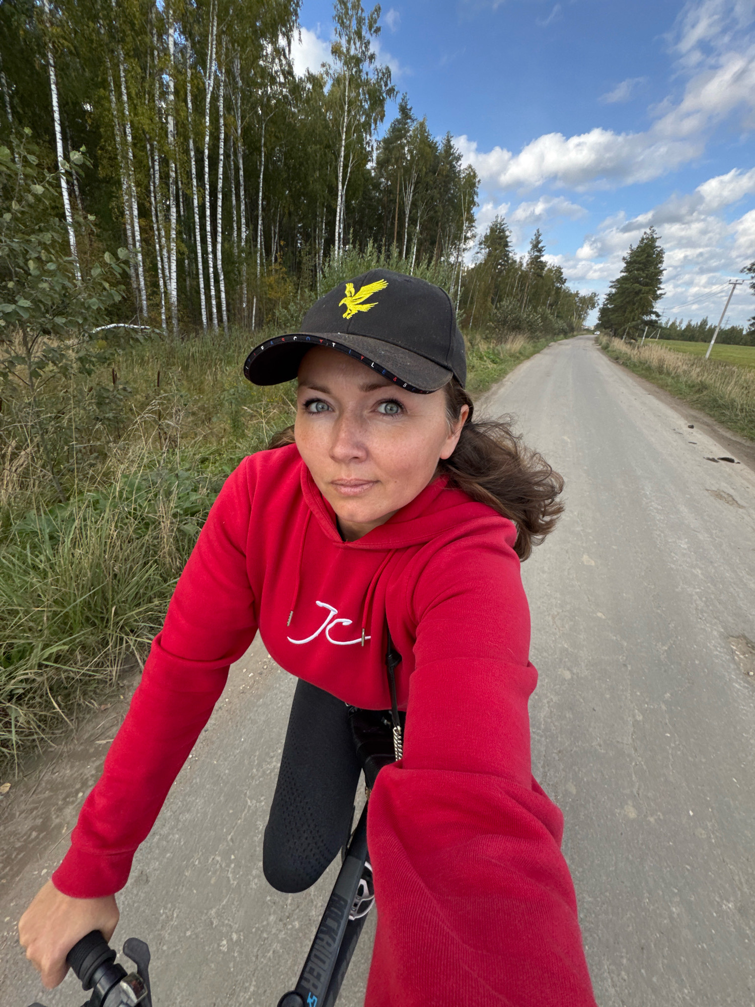 Innolovo, Russia. A woman is riding a bicycle on a dirt road. She is wearing a red hooded sweatshirt, black pants, and a black baseball cap with a yellow eagle logo. She has brown hair and blue eyes. The road is lined with trees and tall grass. The sky is blue with white clouds. The image appears to be a selfie taken by the woman.