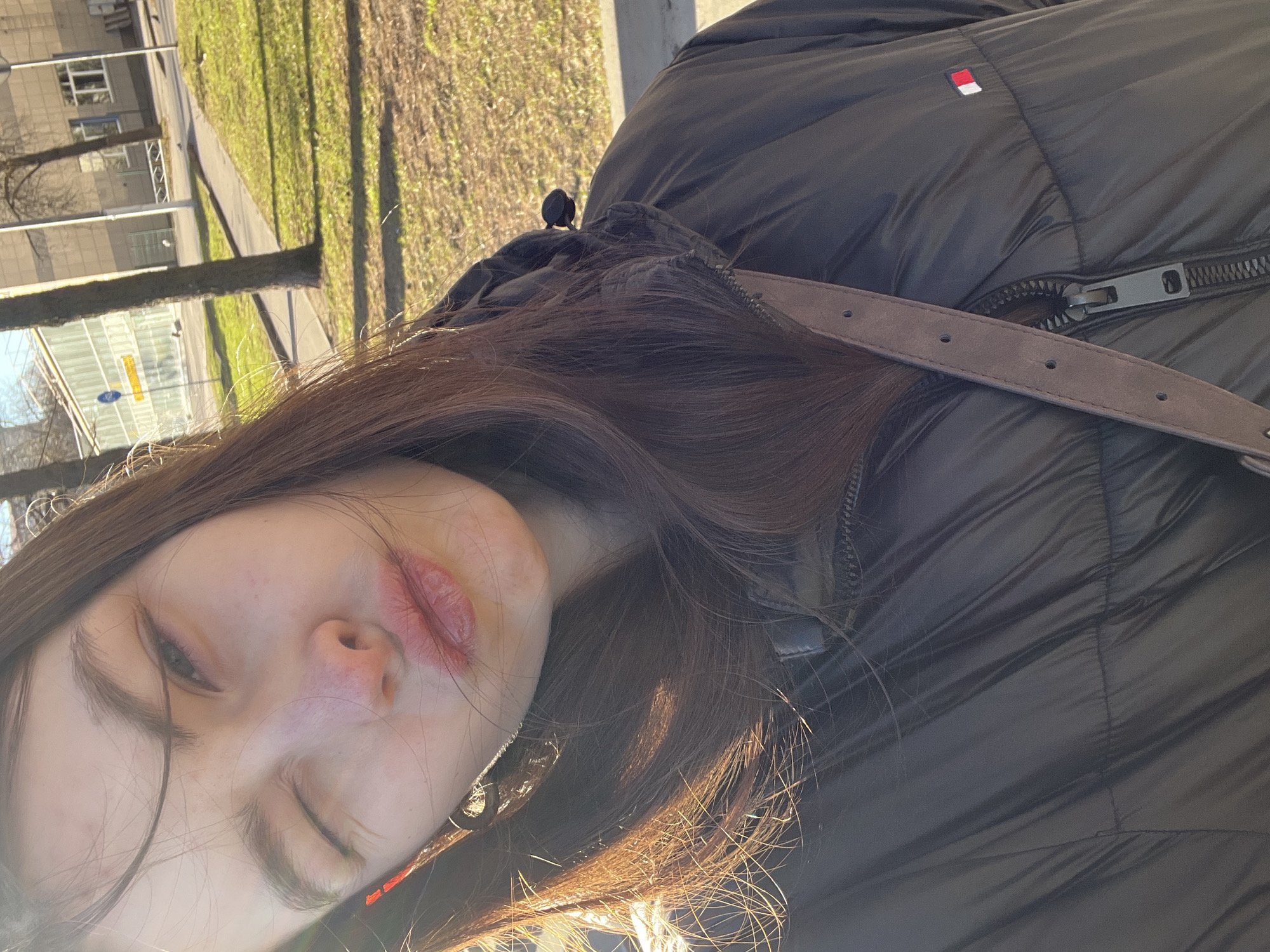 A young woman with long brown hair and fair skin is facing the camera. She has chapped lips, and a Tommy Hilfiger black puffer jacket. She is wearing a brown leather strap around her shoulder. In the background, there is a blurred cityscape of Saint Petersburg, Russia. Trees without leaves are visible, and the sky is bright. A building with multiple windows can be seen. There are patches of grass and some autumn leaves on the ground.