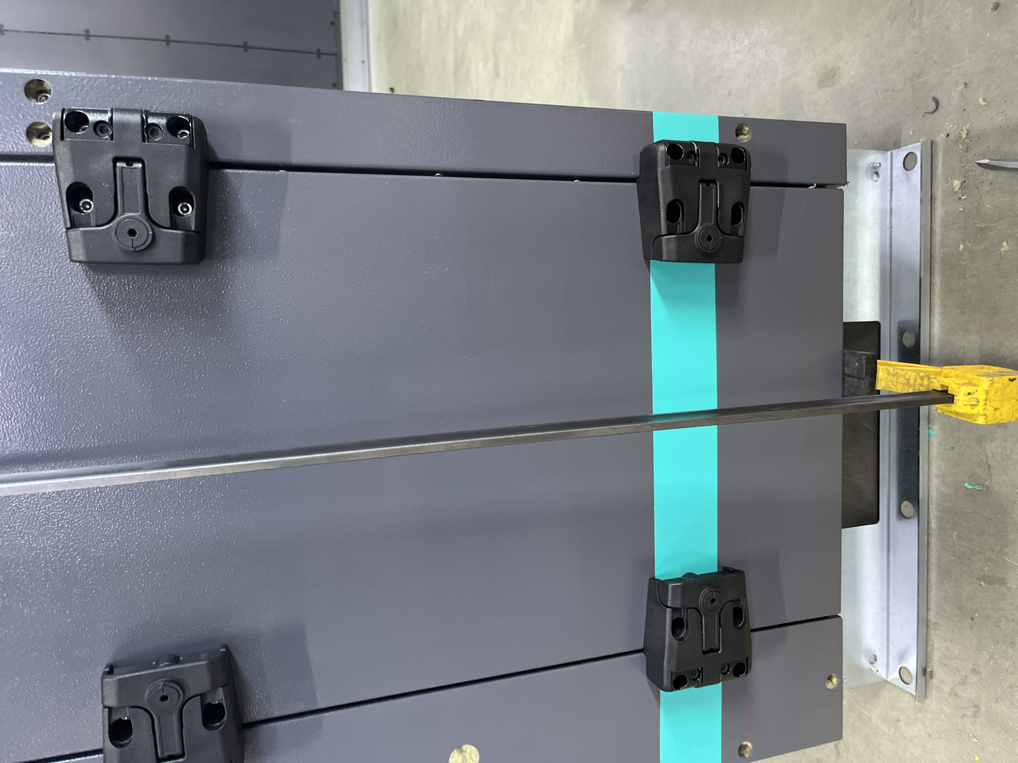 The image shows a close-up view of a dark gray metal cabinet or enclosure, possibly part of industrial equipment. Two black plastic hinges are visible on the left side of the cabinet, one higher up and one lower down. A vertical metal rod is positioned in front of the cabinet, extending from top to bottom, with a yellow handle near the bottom. A turquoise band runs horizontally across the lower portion of the cabinet. The cabinet is mounted on a silver metal base. The background appears to be a concrete floor. No people or specific location details are discernible in the image.
