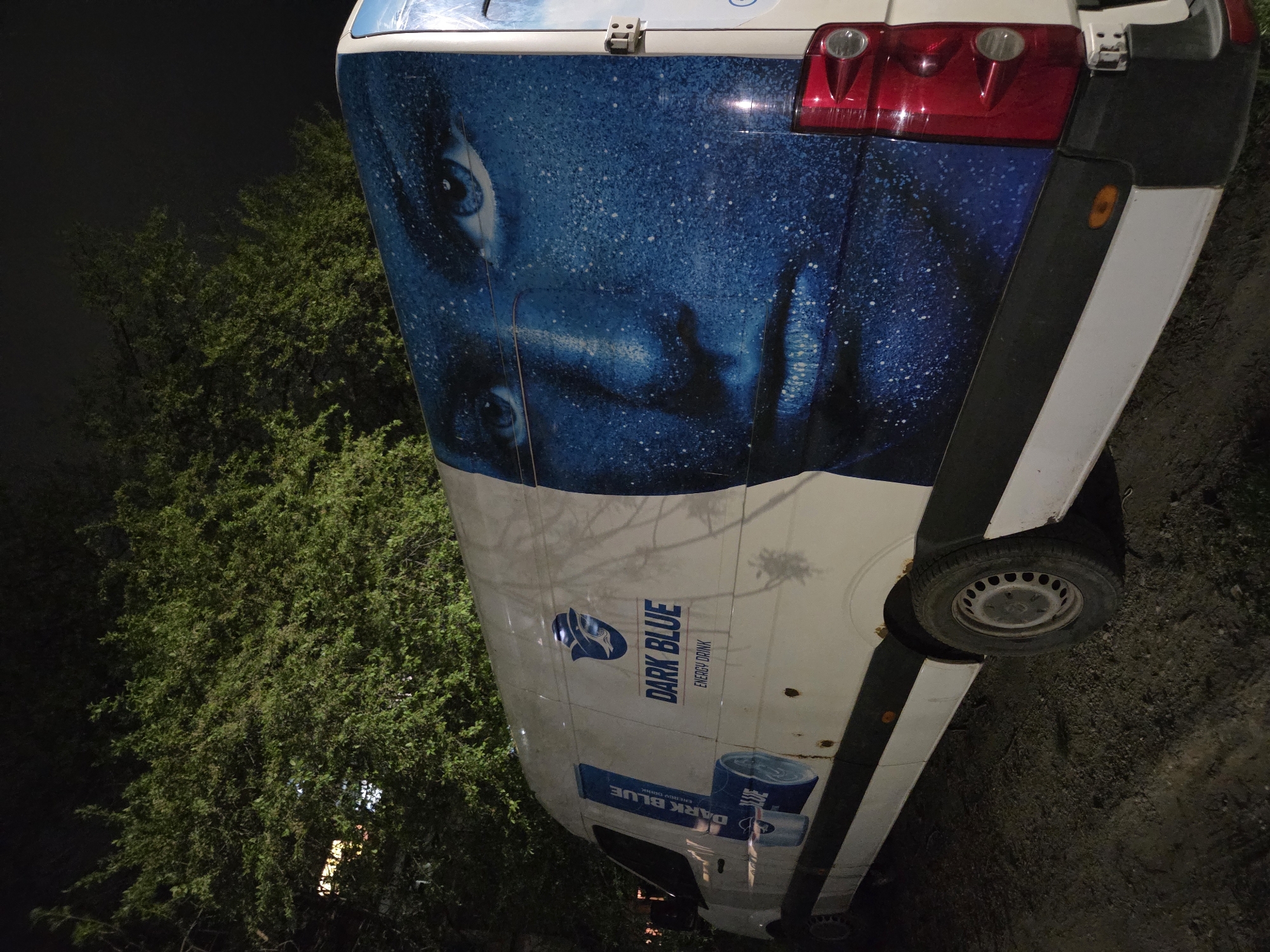 The image depicts the rear of a white van parked on what appears to be a street in Pleven, Bulgaria. The rear of the van is adorned with a large blue advertisement featuring a face and the words "DARK BLUE ENERGY DRINK." There are trees to the left of the van. The lighting suggests it is nighttime. The ground is visible near the right side of the vehicle and a tire is visible.