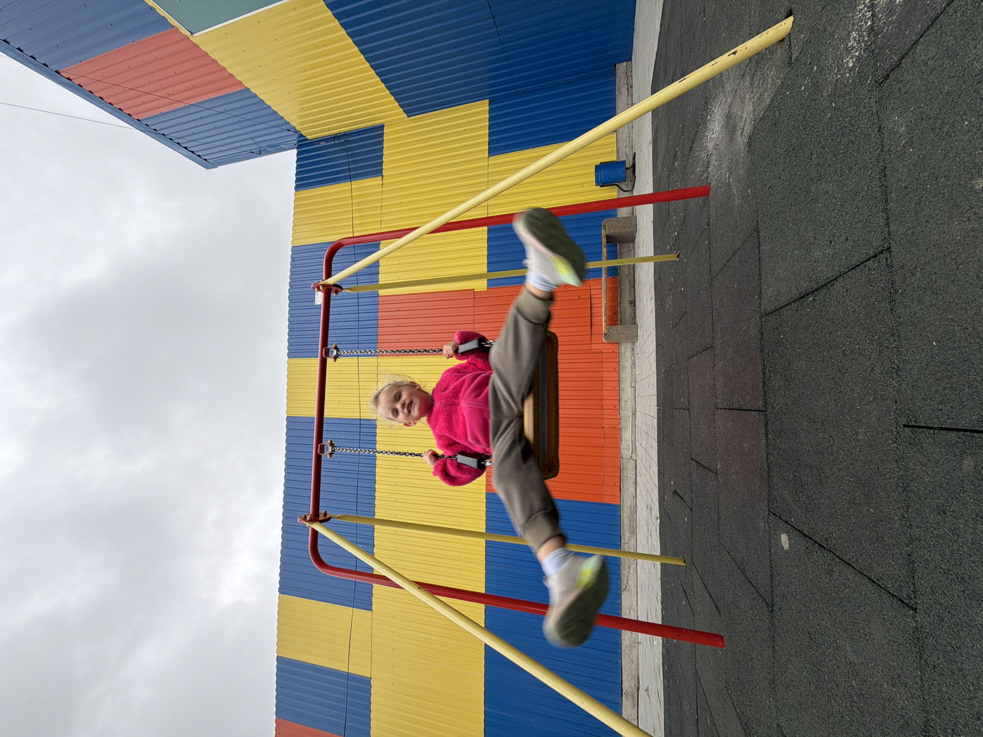 A young child, appearing to be a girl, is captured mid-swing on a playground swing set under an overcast sky in what appears to be an urban playground in Chaikovsky, Russia. The child, wearing a bright pink long-sleeved top, dark grey trousers, and light-colored sneakers with multi-colored soles, is smiling and looking directly at the viewer. Her legs are extended forward and outward, blurred due to motion, indicating active swinging. She holds onto the swing chains with both hands.
The swing set features a red horizontal top bar and yellow diagonal support poles, all anchored into a dark grey rubber safety tile surface on the ground. Beyond the rubber tiles, a lighter grey paved area is visible. In the background, a large building with corrugated metal siding is painted in bold, geometric patterns of blue and yellow, with sections of red and a hint of light green on the far right. The patterns appear as large, interlocking block shapes. To the right of the swing set, a concrete bench and a blue cylindrical object (possibly a trash bin) are visible on the paved area, against the brightly colored building wall. A thin wire or cable is faintly visible traversing the upper right quadrant of the sky.