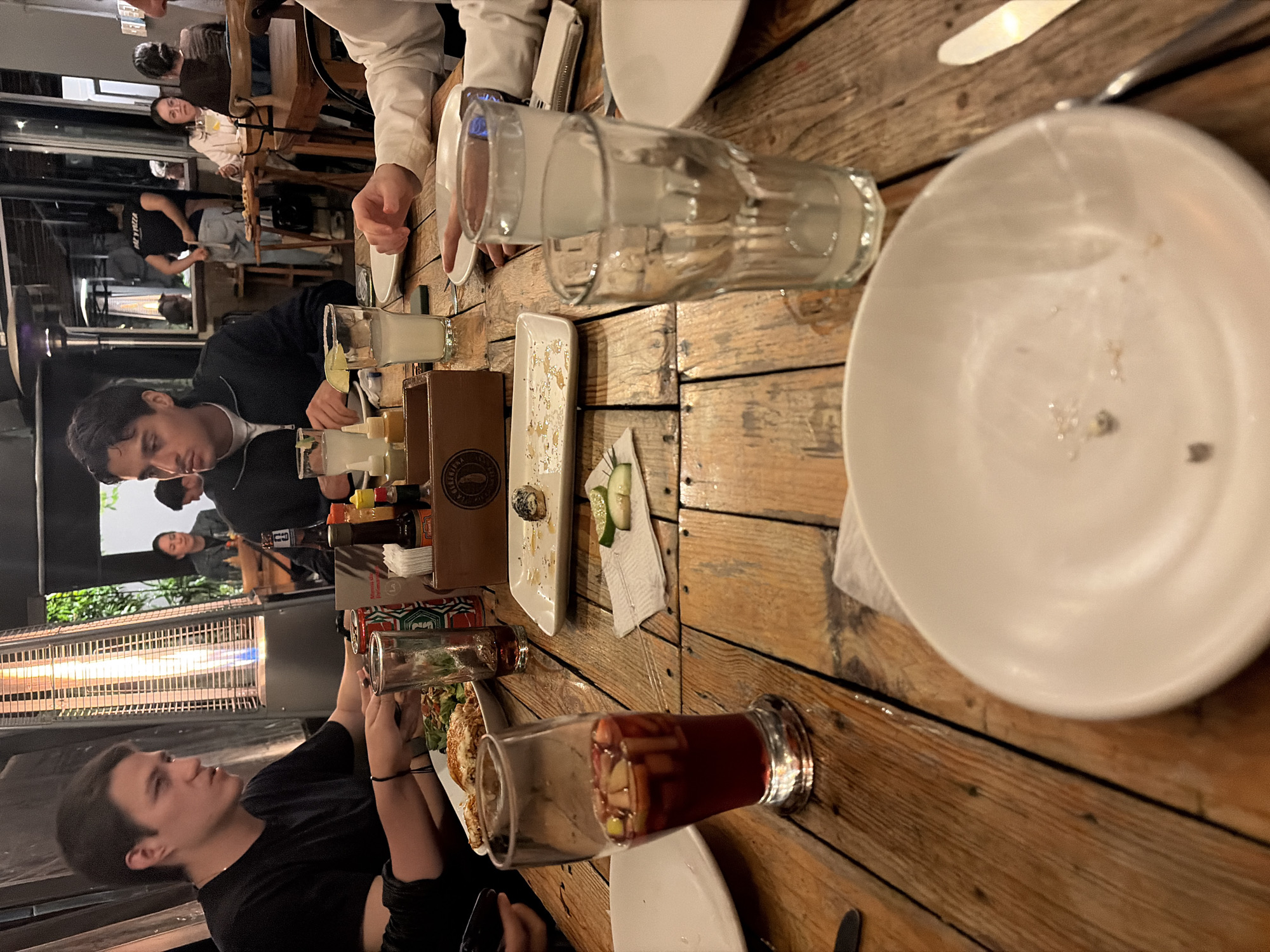 A dimly lit outdoor patio scene at an establishment in an unknown city is captured in this photograph. The focus is on a wooden table laden with remnants of a meal, including empty plates, glassware, and assorted food containers. A few individuals are seen interacting around the table. A person wearing a black top looks directly into the camera, and an individual in a black jacket gazes down at the table, possibly engaged in conversation. In the background, more patrons are visible, seated at other tables in the outdoor space. A tall, illuminated heating apparatus stands near the table, suggesting it might be a cooler evening. The overall impression is that of a casual gathering at an outdoor dining spot.