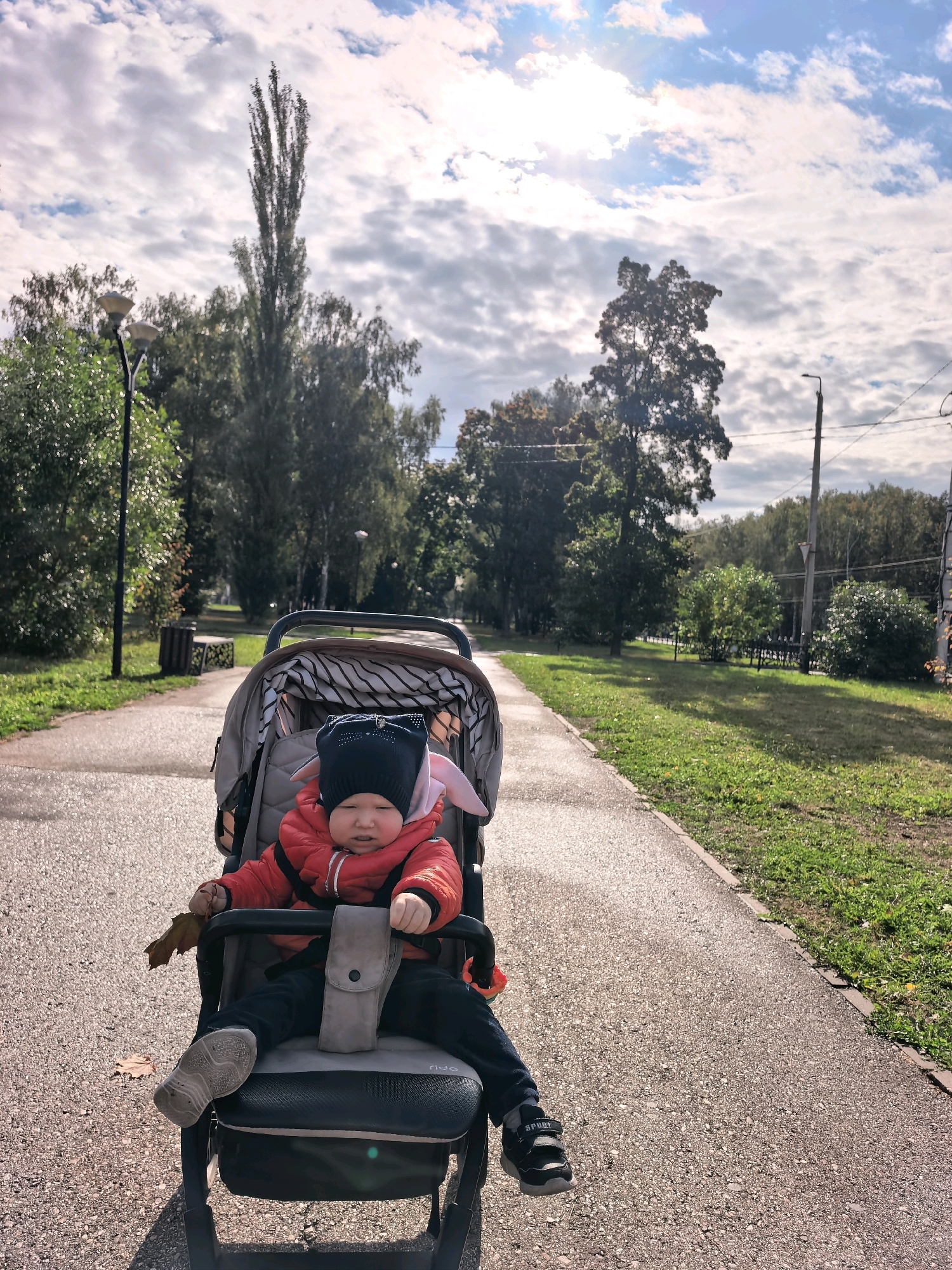 A child wearing a red jacket and a black hat with cat ears sits in a stroller on a paved path in a park. The child is holding a brown leaf in their left hand. The stroller has a grey fabric interior with black and white stripes. To the left of the path, there is a green grassy area with trees and a trash can. A lamppost is visible in the background. To the right of the path, more trees and grass are present, along with a utility pole and wires. The sky is partly cloudy with sunlight visible. The scene appears to be in an outdoor urban park in Sterlitamak, Russia.