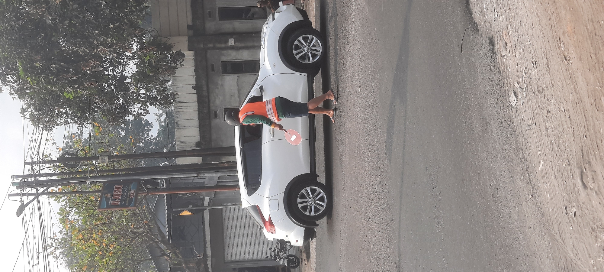 A person wearing an orange vest and a dark helmet stands on a paved road in Siraman, Indonesia, holding a red paddle. The person is walking towards the right side of the image, facing away from the camera. A white car is parked to the left of the person. In the background, there is a building with several windows and a tiled roof. A sign on a utility pole reads "AL-AQSHO" and "4.4 KM". Trees with green and yellow leaves are visible behind the utility pole. The sky is overcast.
