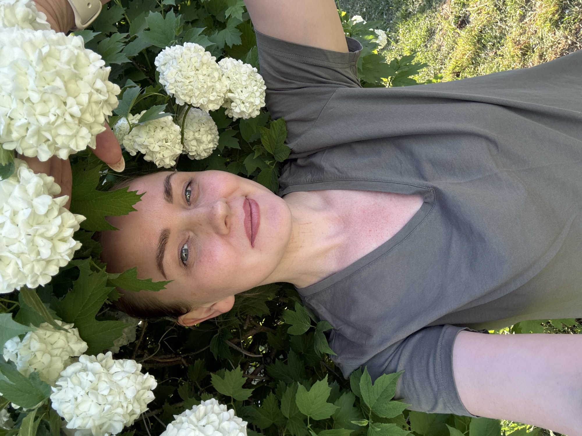 A light-skinned person with brown hair is surrounded by green leaves and white pompom-shaped flowers. The person is wearing an olive-colored shirt. The background appears to be grassy. The location is indicated as Chaikovsky, Russia, but no landmarks or objects in the image confirm this location.
