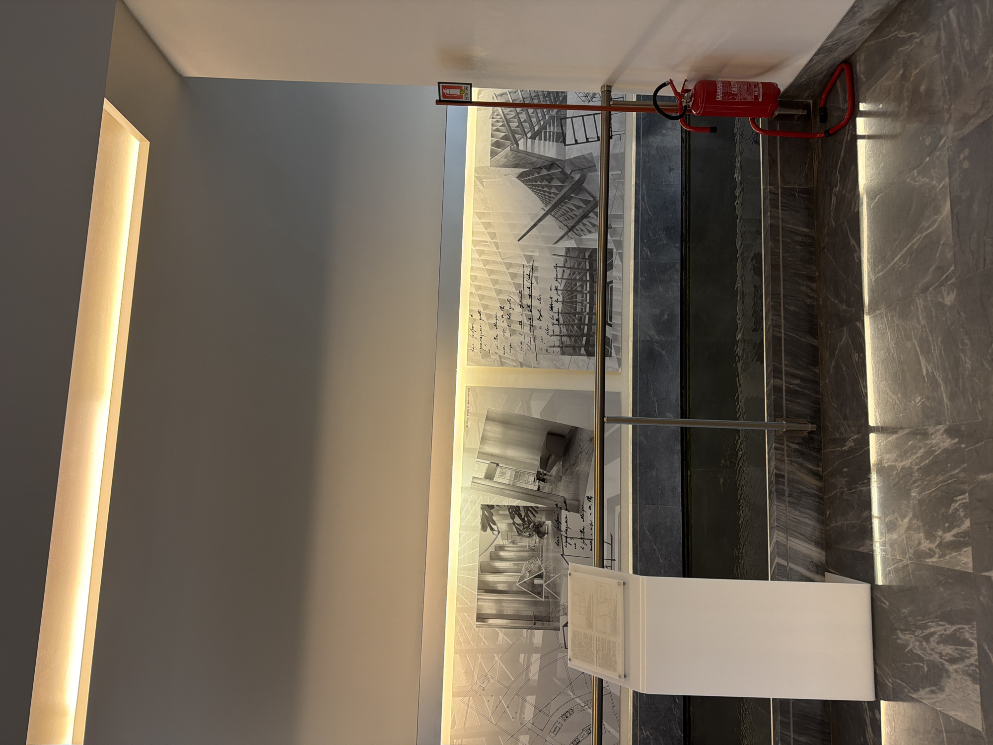 The image captures an interior architectural space, likely a hallway or a landing next to a staircase, featuring a contemporary design and an exhibition display. The scene is illuminated by artificial light, suggesting either indoor conditions regardless of the time of day, or a brightly lit interior.
On the left, a plain grey wall features a long, vertical recessed light fixture, casting a warm, bright glow along its length. This wall forms a backdrop to the primary feature of the scene: large display panels that showcase architectural works. These panels are filled with detailed architectural drawings, possibly blueprints or schematics, overlaid with handwritten script. Interspersed among the drawings are several black and white photographs depicting interior spaces or structural designs. One photo clearly shows the text "GIO PONTI ARCHIVES," indicating a connection to the famous Italian architect Gio Ponti.
A sleek metal railing runs in front of these display panels, guiding visitors along what appears to be a walking path or the edge of a staircase. Adjacent to the railing, a white, rectangular stand with a transparent, angled top presents printed text, likely an informational plaque for the exhibition, though the text is too small to read in detail.
To the right, the floor and a portion of the wall are covered in dark grey marble, characterized by lighter, intricate veining. This surface also features a long, vertical strip of recessed lighting, mirroring the design on the left wall and creating a sense of symmetry and modern elegance. Mounted high on this marble wall is a bright red fire extinguisher, secured in a matching red bracket. The word "ESTINTORE," Italian for fire extinguisher, is clearly visible on its label.
The space conveys a refined and informational atmosphere, indicative of a gallery, museum, or a modern public building, aligning with the provided location context of Milan, Italy. No people are present in the image.