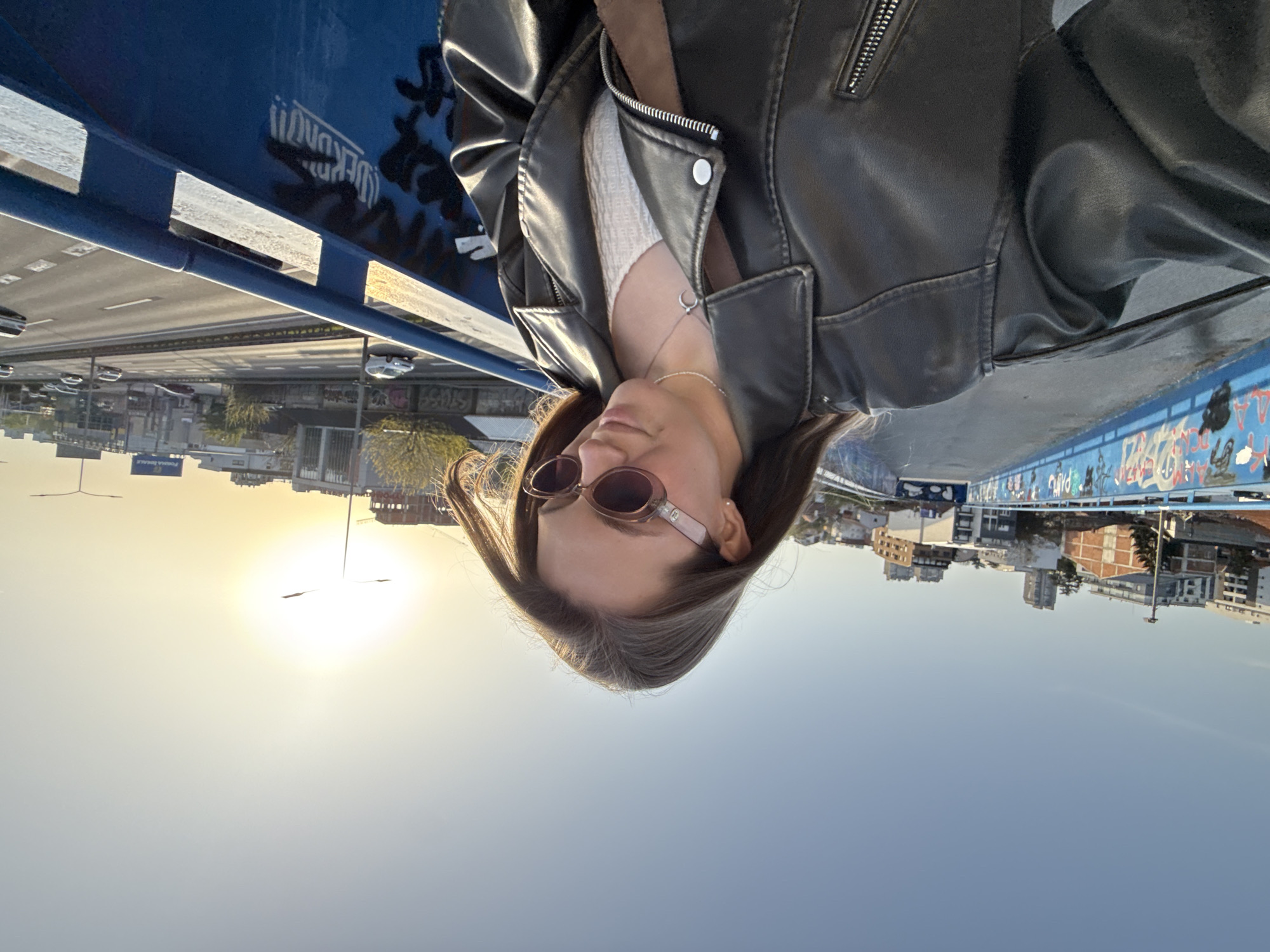 A person stands on the Gazela bridge in Vozdovac Urban Municipality, Serbia. The individual wears a leather jacket and sunglasses. The background shows the cityscape. The sun is visible in the sky.