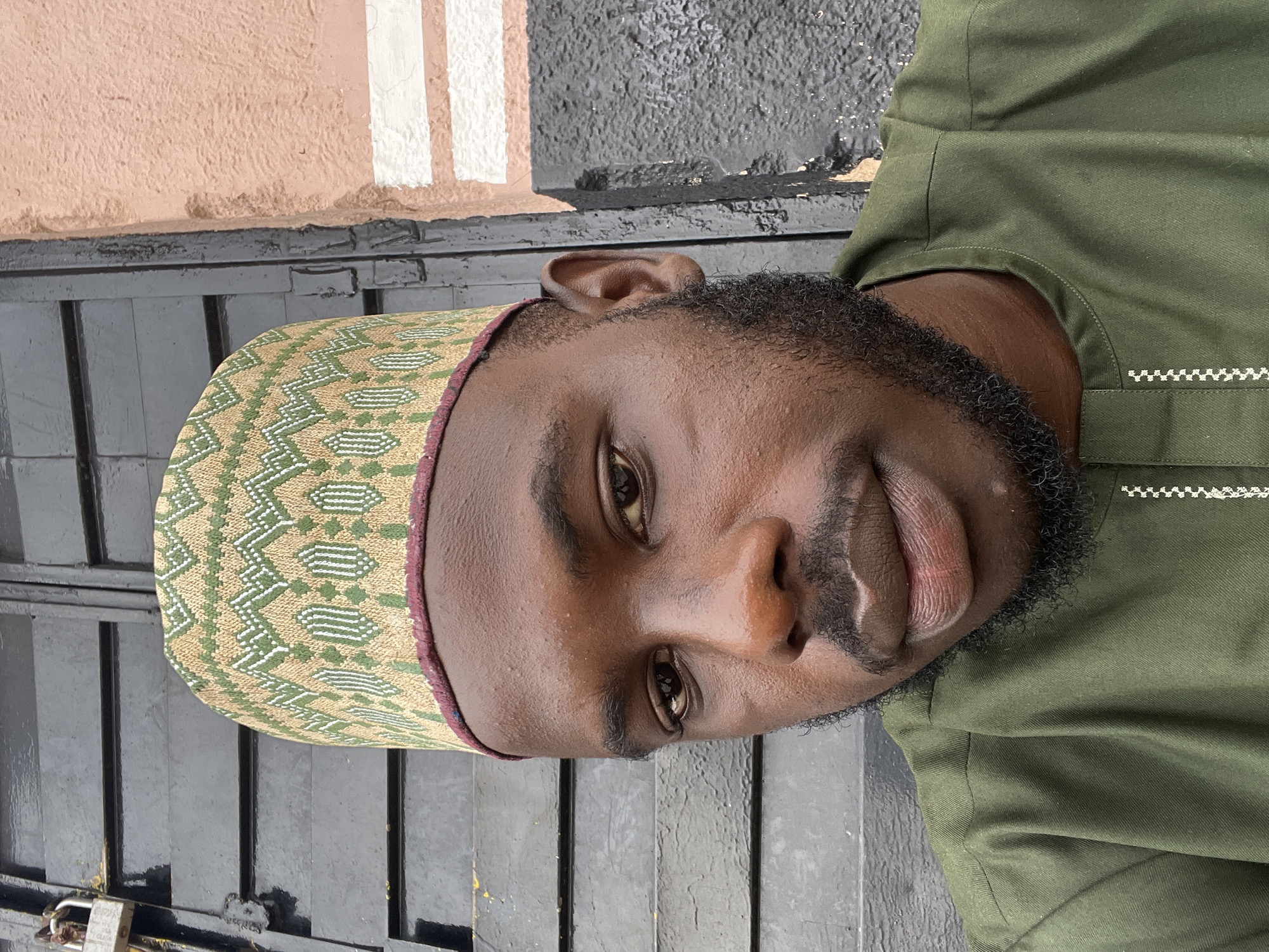A man with dark skin, a beard, and wearing a patterned cap and green garment is shown. He is positioned on a black set of steps near a building with tan and white walls. The location is Gura Top, Nigeria.