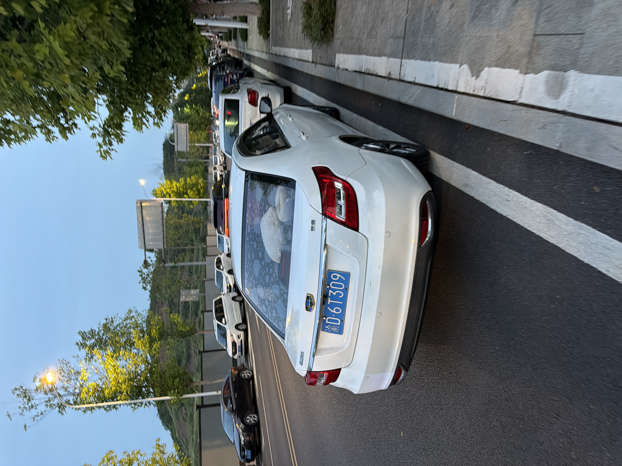 The image shows a white sedan parked on the side of a road in Jiangjin, China, with its license plate reading "D·6T309". The car is facing away from the camera, and its rear bumper and taillights are visible. The road is paved and has white lane markings. In the background, there are other cars, trees, and a hillside. The sky is overcast, and the lighting suggests it is either early morning or late evening. The car's interior is partially visible through the rear window, and appears to contain some luggage or items. The overall scene depicts a typical urban street environment.