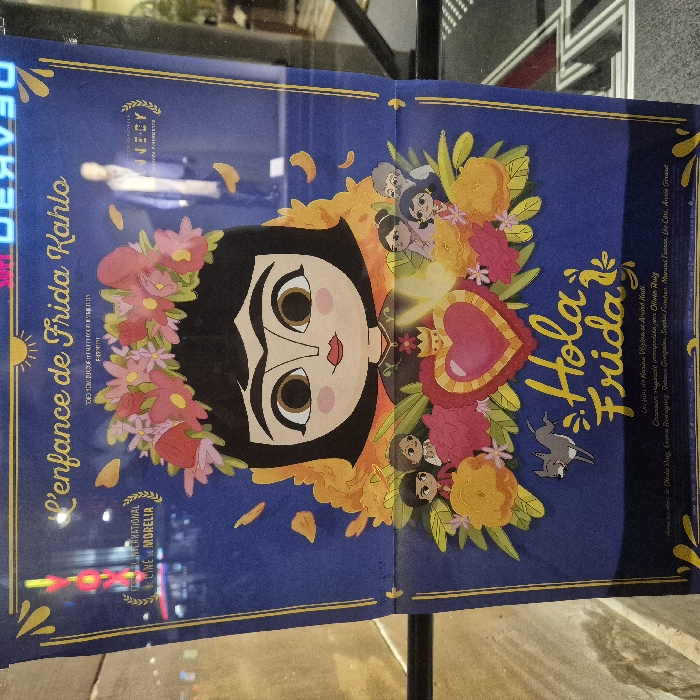 The image showcases a poster for "L'enfance de Frida Kahlo," internationally known as "Hola Frida." The poster features an animated representation of Frida Kahlo with a flower crown. Bright yellow and orange flowers surround her, and several small cartoon figures adorn the scene. "Hola Frida" is written in a playful, golden font. Credits include "Une film de Karine Vezina et Arime Khati," and the poster mentions the Cine de Morelia international film festival.