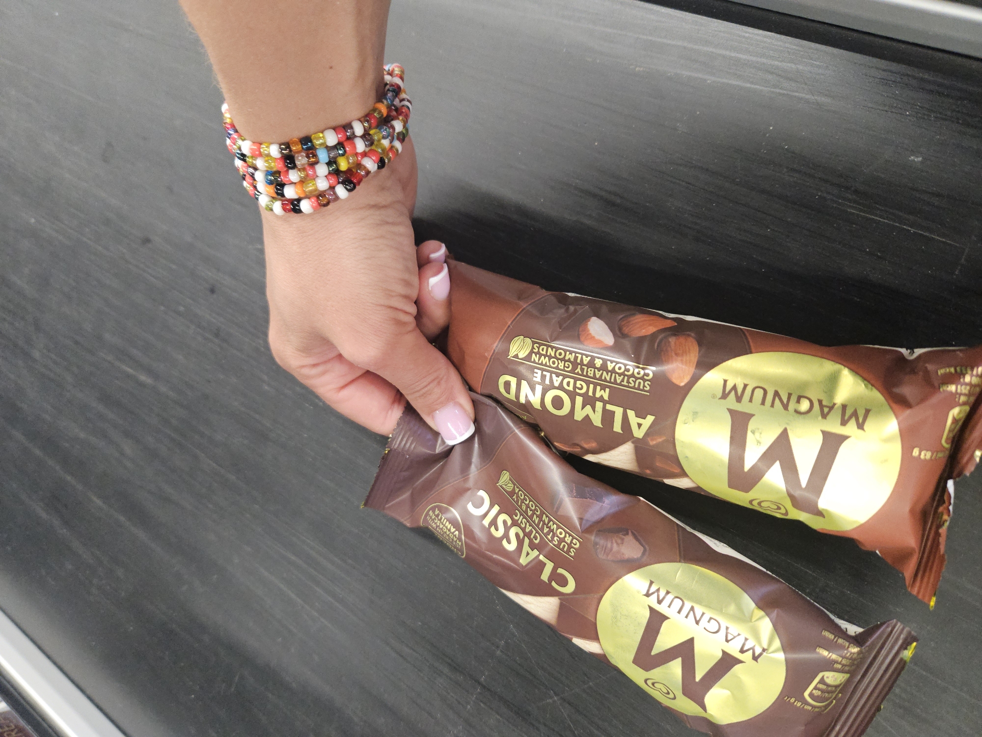 A close-up, high-angle shot captures a person's hand and lower arm holding two Magnum ice cream bars on a dark, possibly black, flat surface, which appears to be a counter. The hand has French-tipped manicured nails and wears a multi-strand, colorful beaded bracelet around the wrist.
One ice cream bar, prominently labeled "MAGNUM ALMOND MIGDALE" with "SUSTAINABLY GROWN COCOA & ALMONDS" and a visual of almonds, is positioned toward the top right. The second bar, held by the hand, is labeled "MAGNUM CLASSIC" and indicates "SUSTAINABLY GROWN COCOA" and "CREAM WITH MADAGASCAN VANILLA." Both wrappers feature the distinctive gold "M" logo for Magnum. Visible text on the packaging also includes nutritional information such as "125 kcal," "205 kcal," and "83 g." The dark surface beneath the items exhibits some light scuff marks or scratches. The activity suggests the person is handling or preparing to purchase the ice cream bars in an indoor, commercial setting.