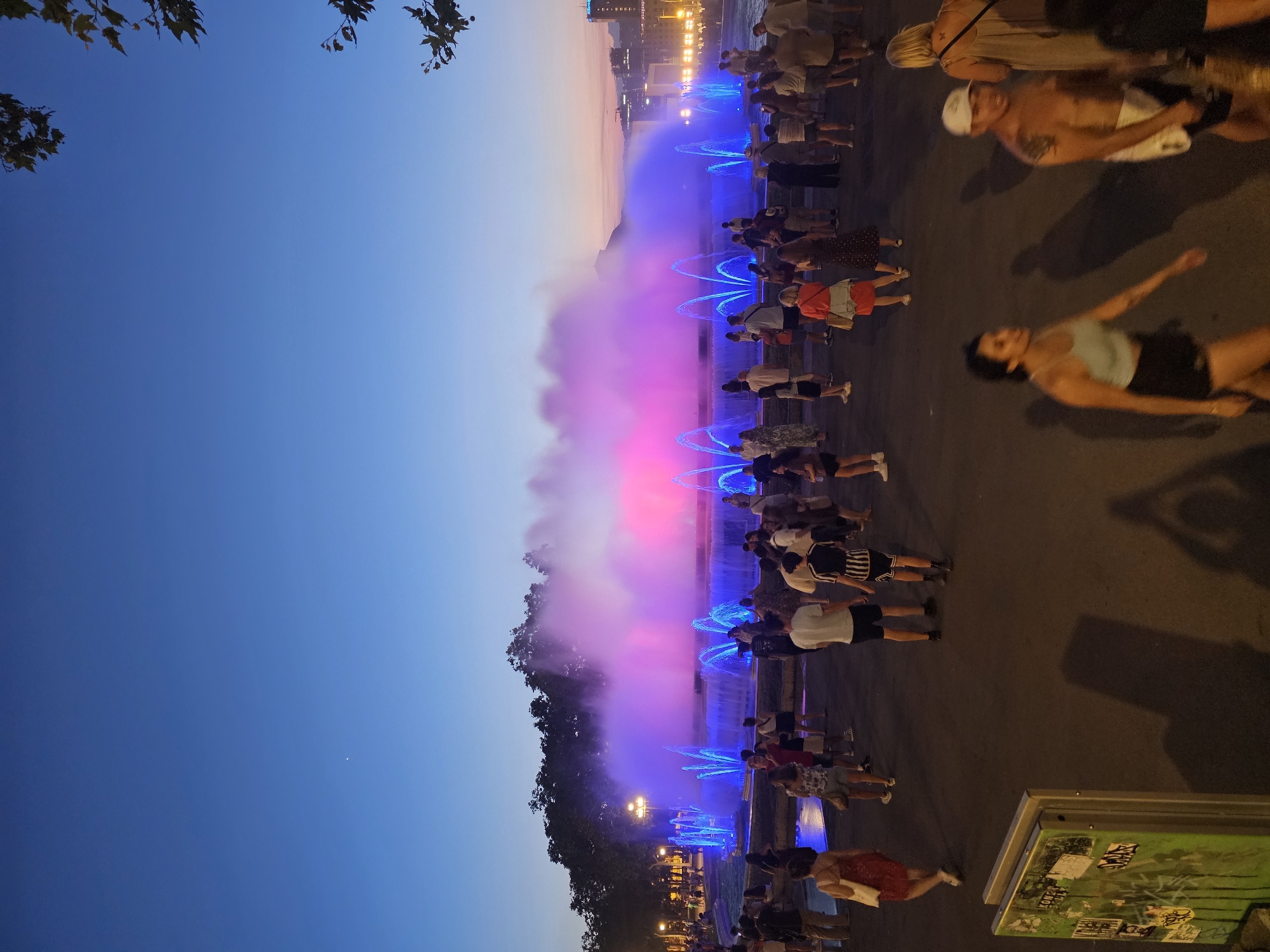 This is a medium-shot, eye-level, outdoor, full-color, documentary-style photograph taken at dusk, featuring the Magic Fountain of Montjuïc in Barcelona, Spain. The fountain is illuminated with vibrant pink and purple lights, creating a dramatic mist. Many people, dressed in casual summer attire, are gathered around the fountain, observing the light show. Some are walking, while others stand and watch. The sky above is a deep blue, transitioning to a lighter hue near the horizon, with faint city lights visible in the distance. Trees line the background behind the fountain, and some branches with leaves are visible in the upper right corner of the frame. In the foreground, a green trash bin covered in graffiti is present. The overall scene captures a lively evening event in a public space.