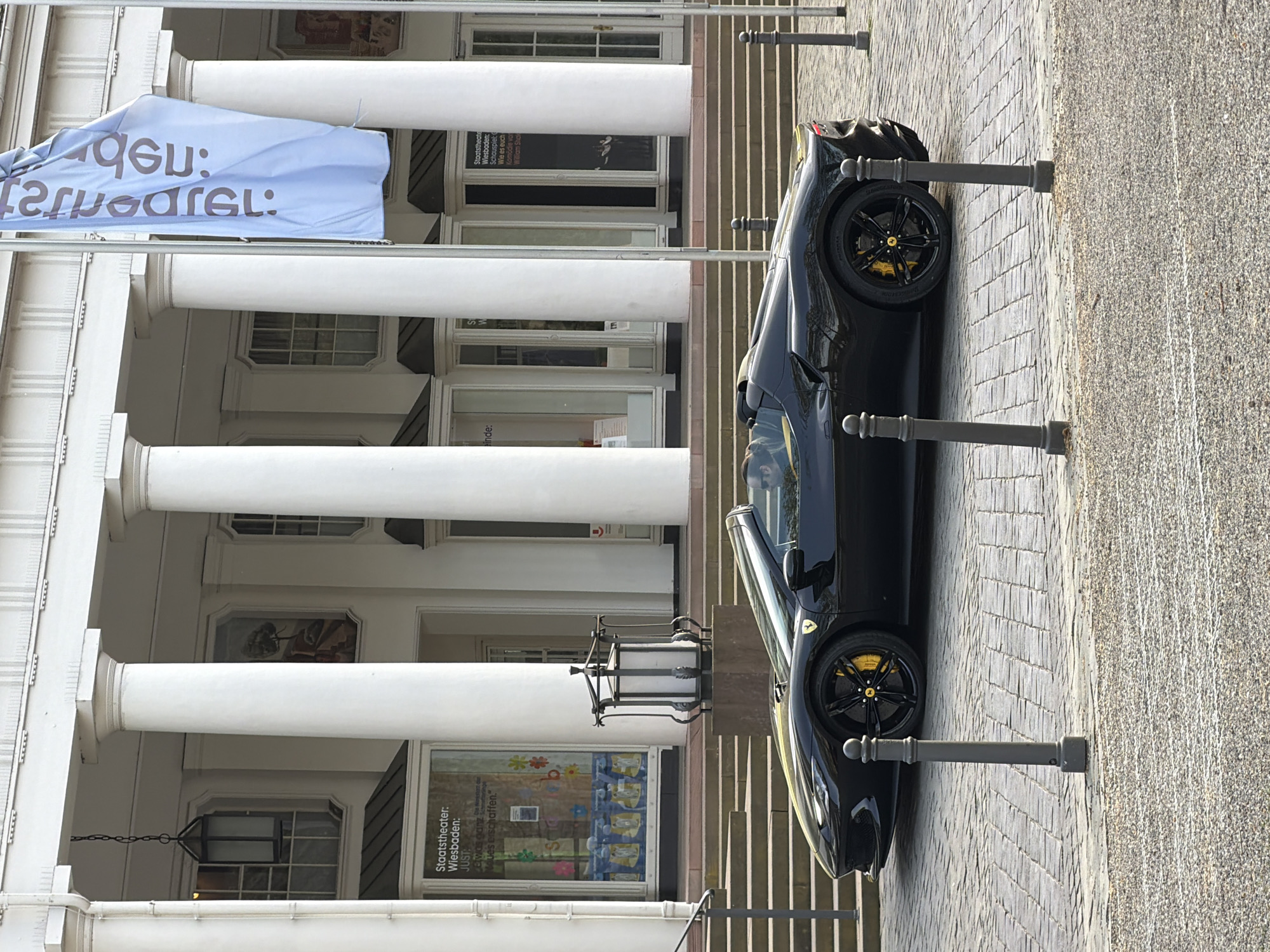 A black Ferrari parked on a brick street in front of a white building with multiple columns is visible. A sign is visible at the top of the building that reads "qGม: Staatstheater: Wiesbaden: JUST: Etwas dan Moue's erschaffen." Another sign is visible below that one, reading "Staatstheate Wiesbaden Schauspiel Wie es euch Komödie vor William Shak." There are metal posts lining the street. The location is likely in the city of Wiesbaden, Germany, based on the text visible on the signs.