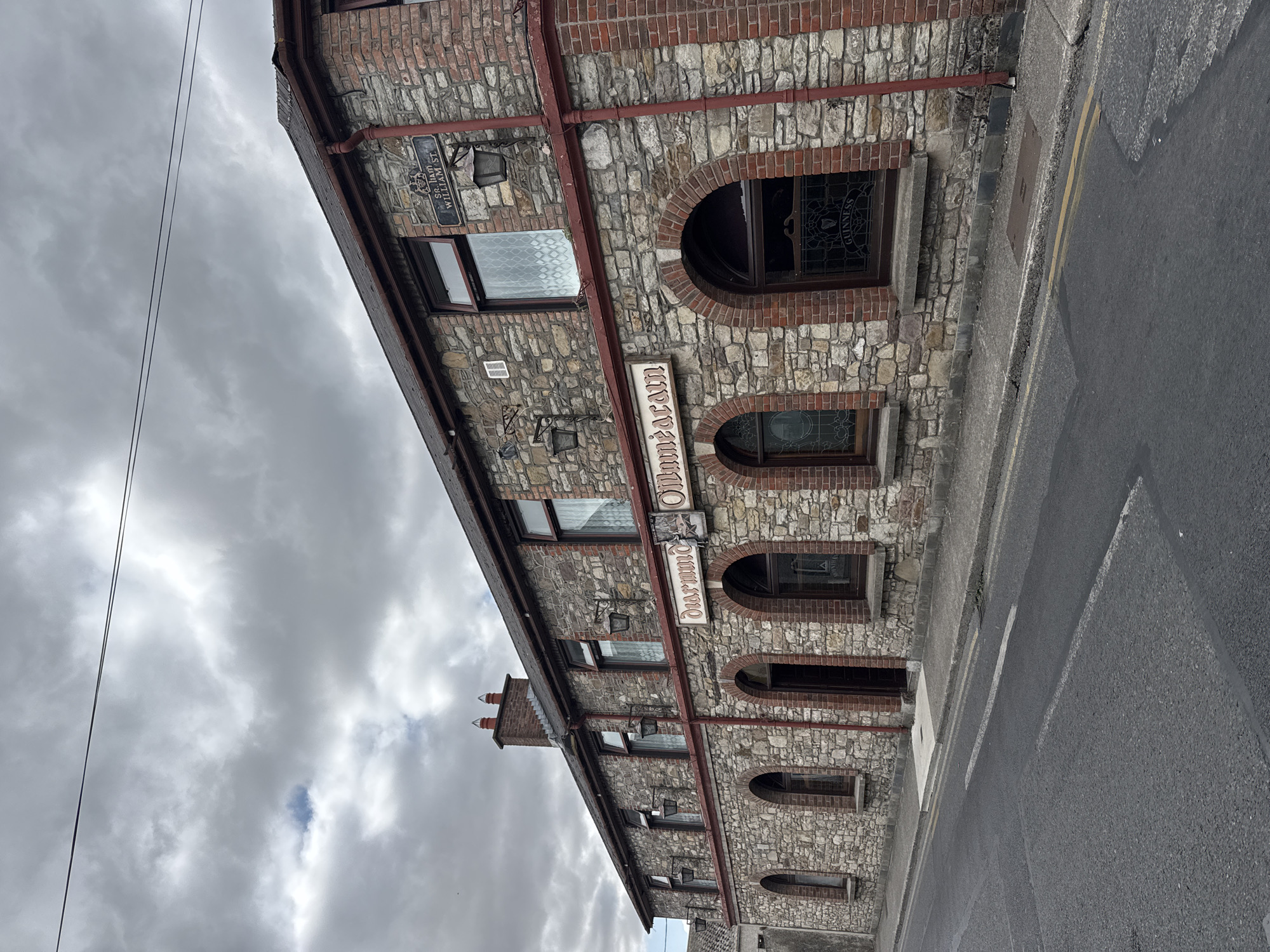 A two-story stone and brick building is depicted under a cloudy sky in Clonmel, Ireland. The building has a red-tiled roof with a red-painted gutter and downspout. The lower facade is constructed of rough-hewn stone blocks, while the upper facade features brickwork. Several arched windows with dark frames are visible on the lower level, some with stained glass. The upper level has rectangular windows with white patterned curtains. A sign above the arched windows reads "Duaramda Ollumdaairn". A street sign on the upper right corner of the building reads "SR. LIAM WILLIAM ST.". Two outdoor lamps are mounted on the building's facade. The building faces a paved street with yellow double lines. The environment appears to be an urban setting, likely a commercial or residential area.