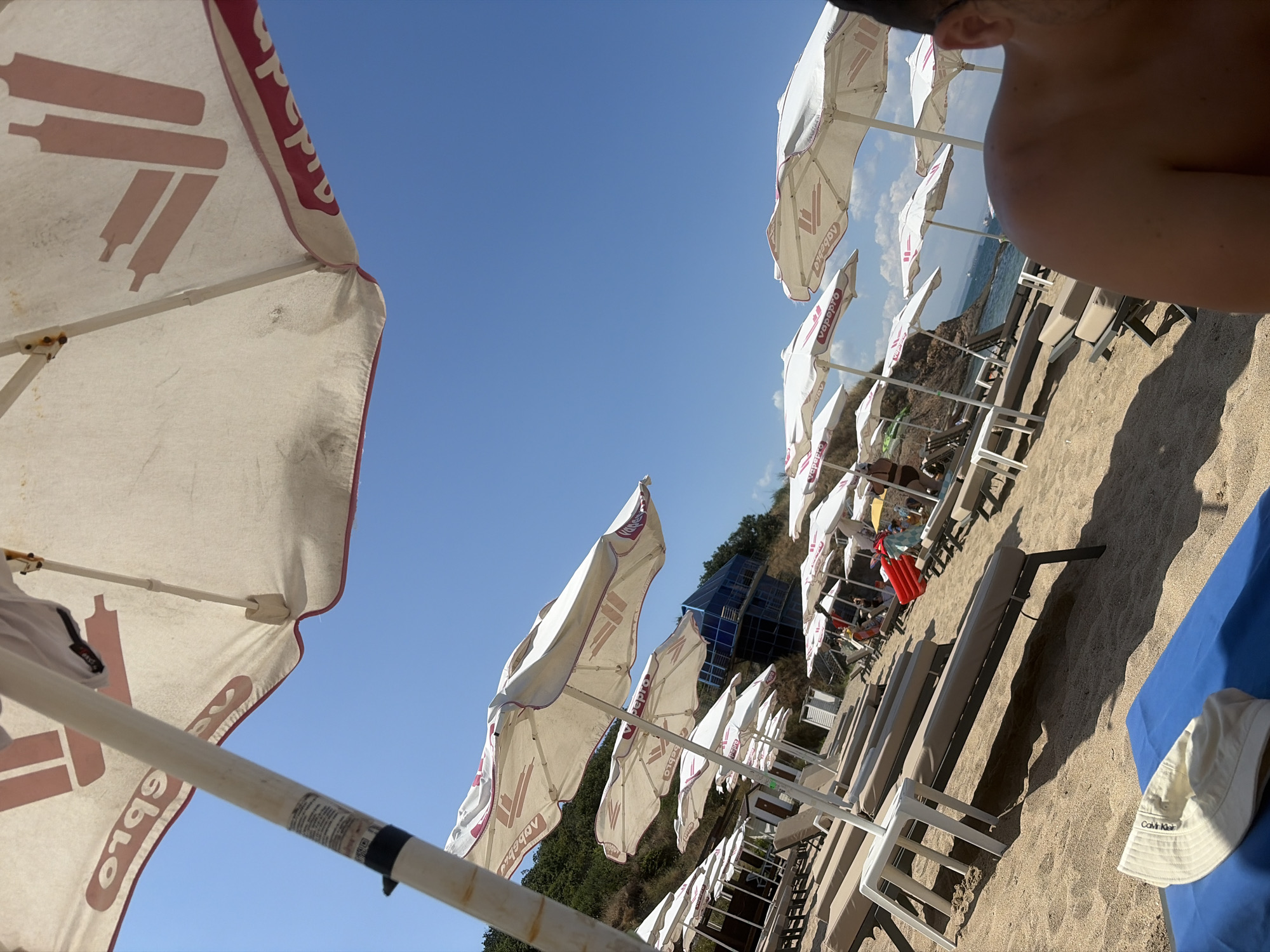 An outdoor, ground-level, eye-level, wide shot captures a sunny beach scene on a clear day. The image shows a row of unoccupied beach chairs and umbrellas set up on a sandy beach. The umbrellas are white with pink lettering and logos. In the background, there is a body of water, some greenery on a hillside, and a building with a blue, angular roof. A person's shoulder and upper back are visible in the lower right corner of the frame, suggesting the photo was taken from their perspective. The overall impression is a serene and inviting beach environment. Based on the visual cues and context, the scene is likely at a beach resort within the city of Burgas, Bulgaria, known for its coastal attractions.