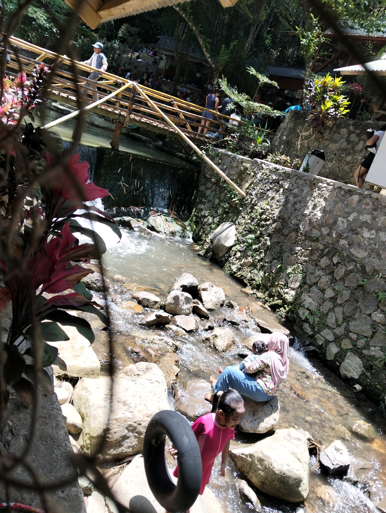 The image depicts a scenic outdoor location, possibly a natural spring or waterfall area, with a focus on a shallow, rocky stream. In the foreground, a young girl in a pink swimsuit is standing in the water, looking towards the viewer with a smile. To her right, a woman wearing a pink hijab and patterned shirt sits on a rock in the stream, holding a child.
The stream flows over rocks and appears to be a clear, shallow body of water. The banks of the stream are lined with stones and vegetation. In the background, a wooden bridge spans the stream, with several people visible walking on it. Some structures, possibly huts or resting areas, are nestled among trees and greenery on higher ground. The environment suggests a recreational or natural tourist spot. While the image provides no explicit confirmation of its location, the mention of "Subang, Indonesia" implies this setting is within that region.
