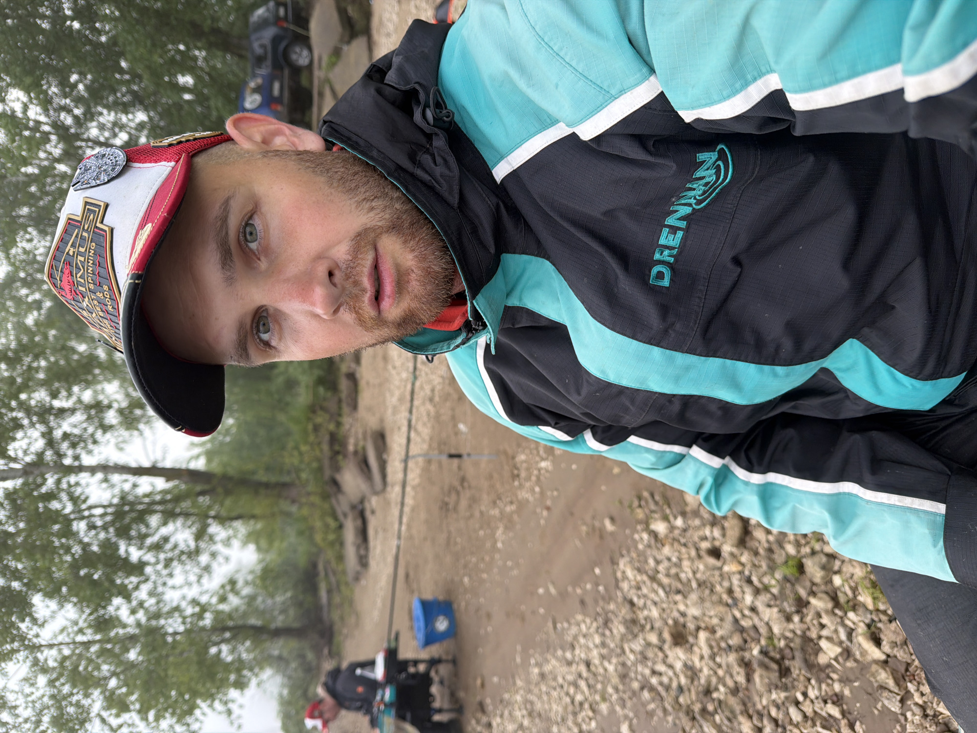 A medium close-up, rotated image captures a man outdoors in Noviy, Russia, looking directly at the viewer with a neutral expression. He has light green eyes and a short beard. He wears a distinctive baseball cap, primarily white, red, and black, featuring the text "TIMIUS," "SPINNING," and "RODS," along with a prominent silver Millennium Falcon pin on its side. His attire includes a black and teal jacket with white stripe accents. The brand "DRENNAN" is visible in teal lettering on the jacket's black section.
The setting appears to be a natural, rugged environment, likely a river or lake bank, characterized by rocky and muddy ground. The sky is overcast, contributing to a muted, diffused lighting. In the background, another person is visible, seated on what appears to be fishing equipment, with a fishing rod extending from their position and a blue bucket nearby, indicating fishing activity. Further in the distance, a dark blue vehicle is partially obscured by trees. The overall scene suggests a leisurely outdoor activity like fishing on a cloudy day.