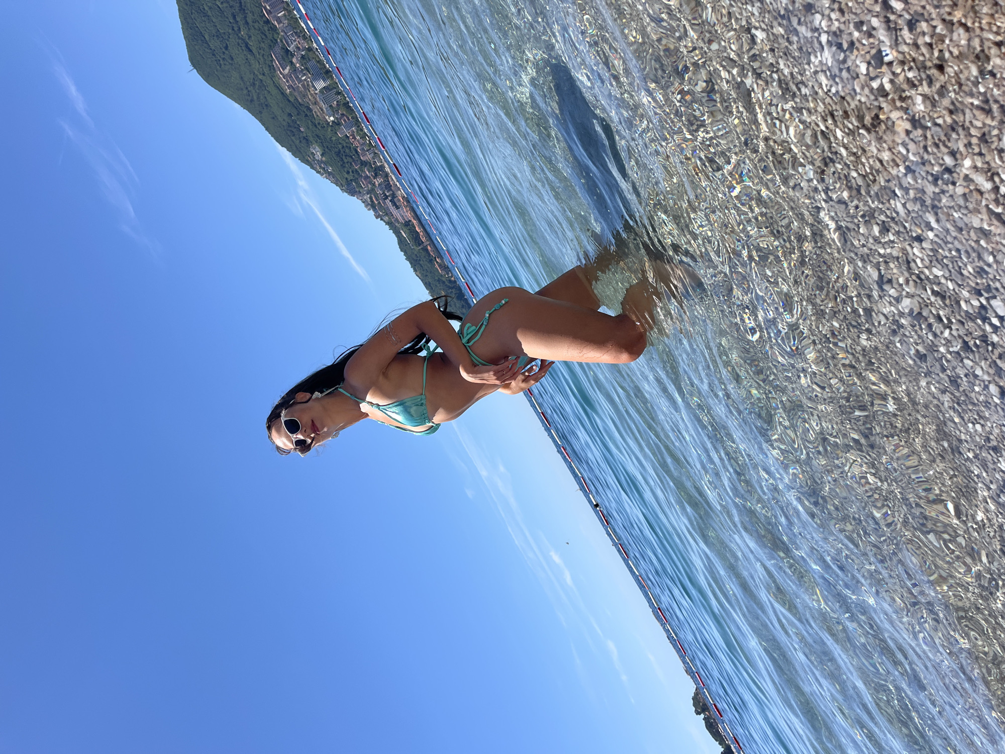 In Budva, Montenegro, a woman stands in shallow, clear turquoise water near a pebble beach. The woman, wearing a green bikini and sunglasses, poses looking over her shoulder. The water is calm with small ripples, and the sun casts light reflections on the surface. In the background, a mountainous coastline dotted with buildings stretches across the horizon under a clear blue sky. Red and white buoys mark a line in the water.
