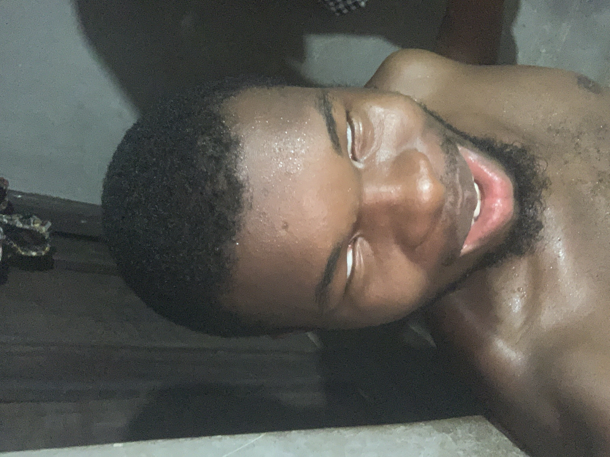This media file presents a close-up, tilted portrait of a man, possibly of African descent. His head is angled, and his eyes are rolled back, showing only the whites. His mouth is open, revealing his tongue and some teeth. His skin, particularly on his face and visible upper chest, is glistening with what appears to be sweat or water. He has short, dark hair and some facial stubble around his chin and jawline.
The man's posture and expression suggest a state of intense physical or emotional experience, which could range from extreme exhaustion or distress to a moment of intense pleasure or altered consciousness.
The setting appears to be dimly lit, likely indoors, with a dark, indistinct background. A faint, lighter-colored surface, possibly concrete or tile, is visible along the bottom edge of the frame. The overall impression is one of rawness and immediacy.
No distinct objects other than the man himself are clearly identifiable. There is no visible text. While the image is provided with the context of being taken in Onitsha, Nigeria, there are no specific visual cues within the image itself to confirm this location. The time of day appears to be low-light, possibly night, or an interior space with minimal illumination.