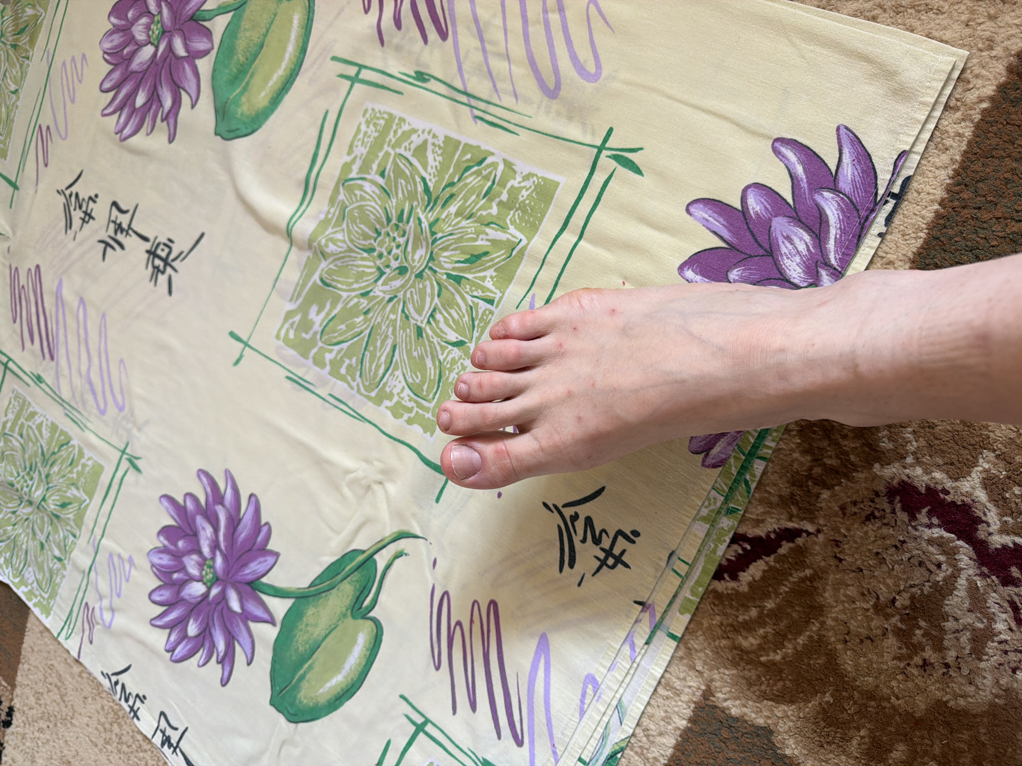 An image shows a bare foot resting on a yellow sheet with a floral pattern of purple and green flowers. There are also some stylized black brushstrokes that appear to be Chinese calligraphy. The sheet is on top of a brown and red patterned carpet. The scene appears to be indoors.