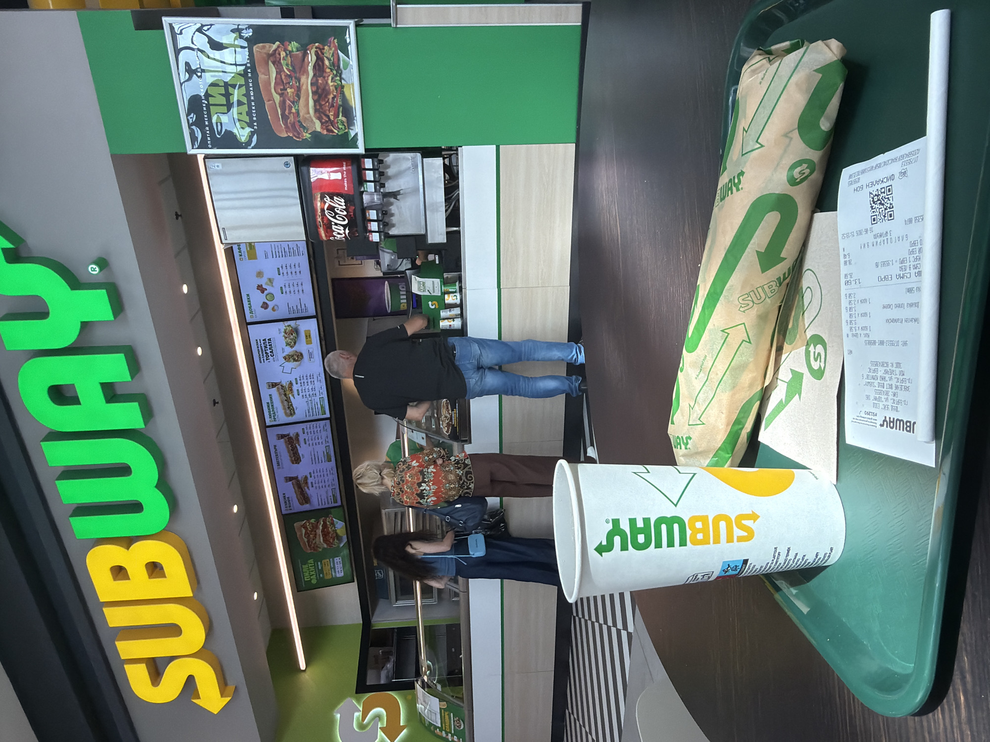 The image depicts the interior of a Subway restaurant located in Burgas, Bulgaria. In the foreground, a green plastic tray holds a Subway paper cup, a wrapped sandwich, and a receipt. The cup features the Subway logo in white, yellow, and green. The sandwich is wrapped in brown paper printed with green Subway branding. The receipt, also with Subway branding, is partially visible.
In the middle ground, several people are visible. Two women stand at the counter, appearing to order food. One woman has long dark hair and wears a blue top and dark pants, holding a blue purse. The other woman wears a patterned colorful blouse and brown pants. A man in a black t-shirt and blue jeans is also at the counter, possibly waiting for his order. Behind the counter, staff members are likely preparing food, and a Coca-Cola dispenser is visible.
The background shows the Subway branding prominently displayed on the wall with illuminated yellow and green letters. Menu boards with images of sandwiches and prices are displayed above the counter. The restaurant has a modern interior design with green and light-colored accents. The floor has a black and white striped pattern.
