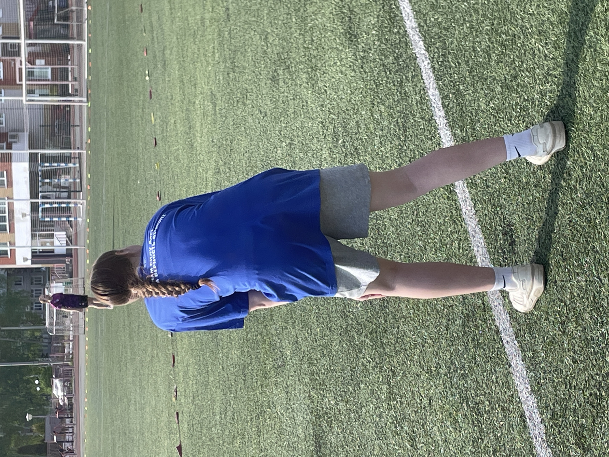 A woman in a blue t-shirt and gray shorts stands on a green artificial turf field in Zav'yalovo, Russia. She has a braid and is wearing white socks and sneakers. A white line is visible on the turf. In the background are buildings, a soccer goal, and another person in the distance.