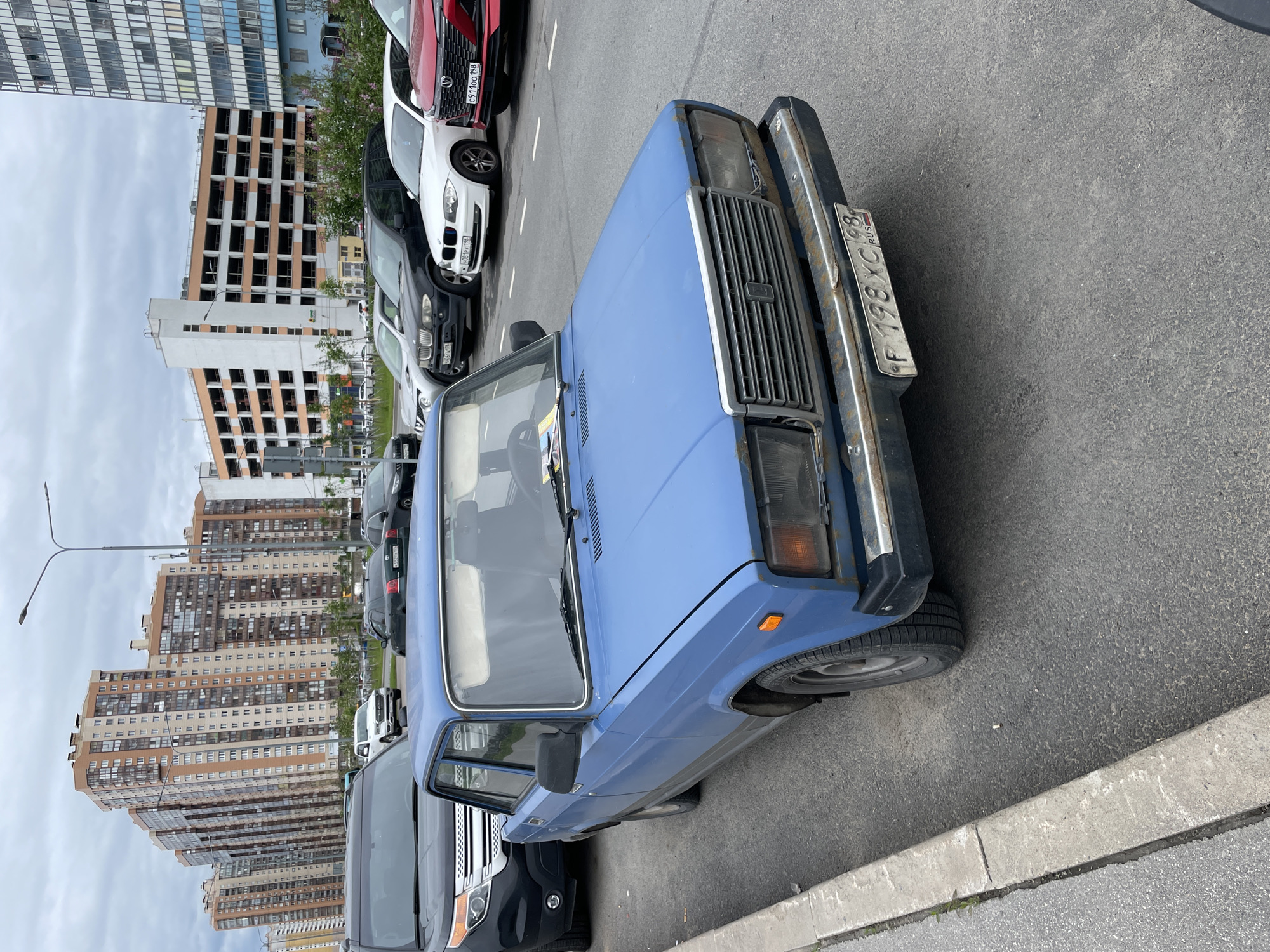 The image shows a blue Lada car parked on an asphalt road next to a concrete curb. The car's license plate, "P 198 XC 98 RUS," indicates it is registered in Russia. Other cars are parked in the background, including a red Changan, white BMW and some SUV. High-rise buildings are visible in the background, suggesting an urban area. The sky is overcast. The location is likely Saint Petersburg, Russia, based on the license plate number indicating the region.