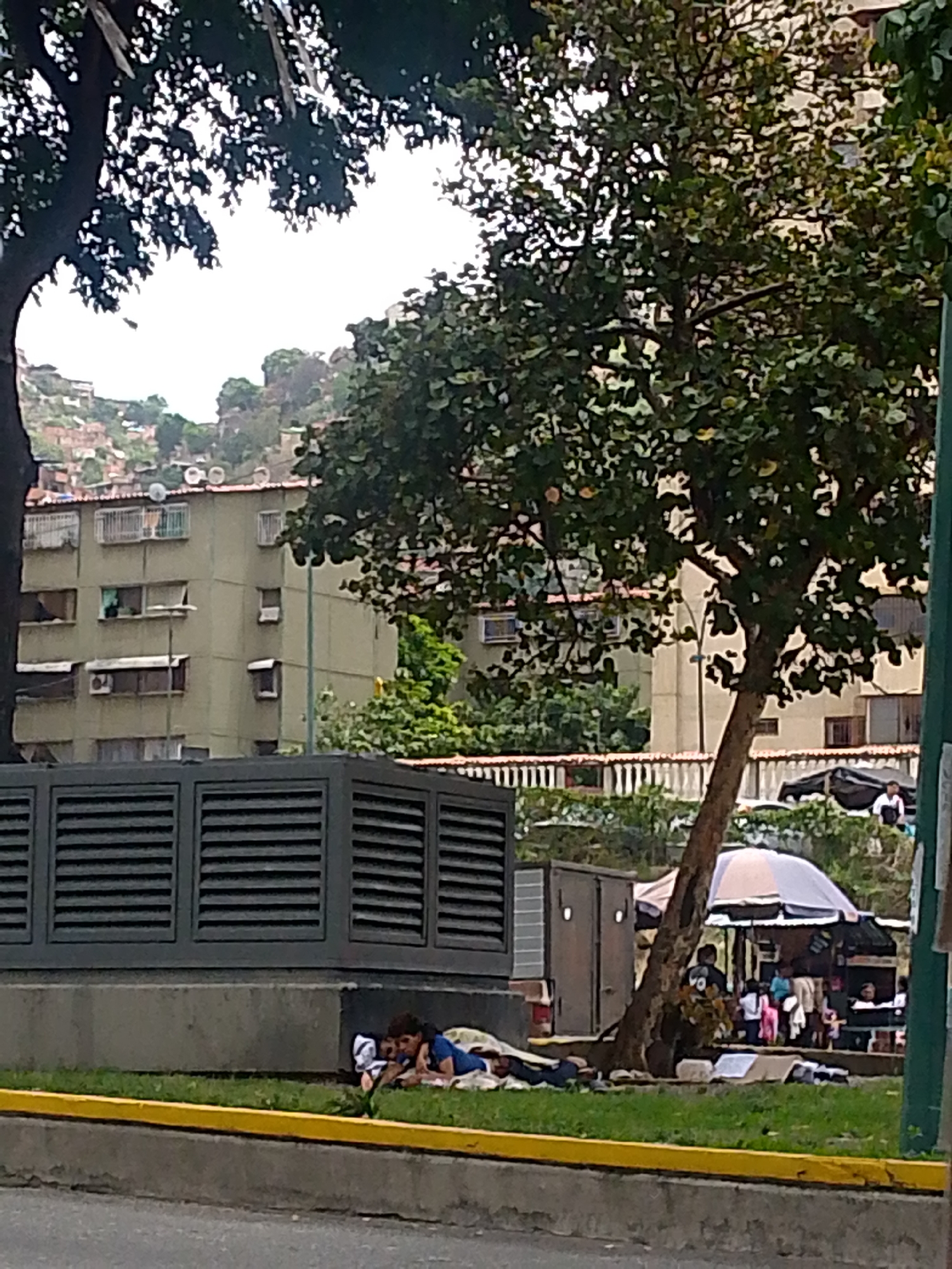 The image shows a scene in Caracas, Venezuela. In the foreground, two people are lying on the grass next to a grey concrete structure with horizontal slats. They appear to be resting or sleeping. Behind them, there's a line of portable stalls or kiosks, possibly selling goods or services. People are visible near the stalls, some standing and some seated. A large tree is situated between the resting individuals and the stalls, with its trunk visible and branches extending outwards. In the background, multi-story buildings are visible, with some showing signs of weathering. A hillside with some visible structures rises in the far background. The lighting suggests an overcast day. A yellow curb marks the edge of the grass.