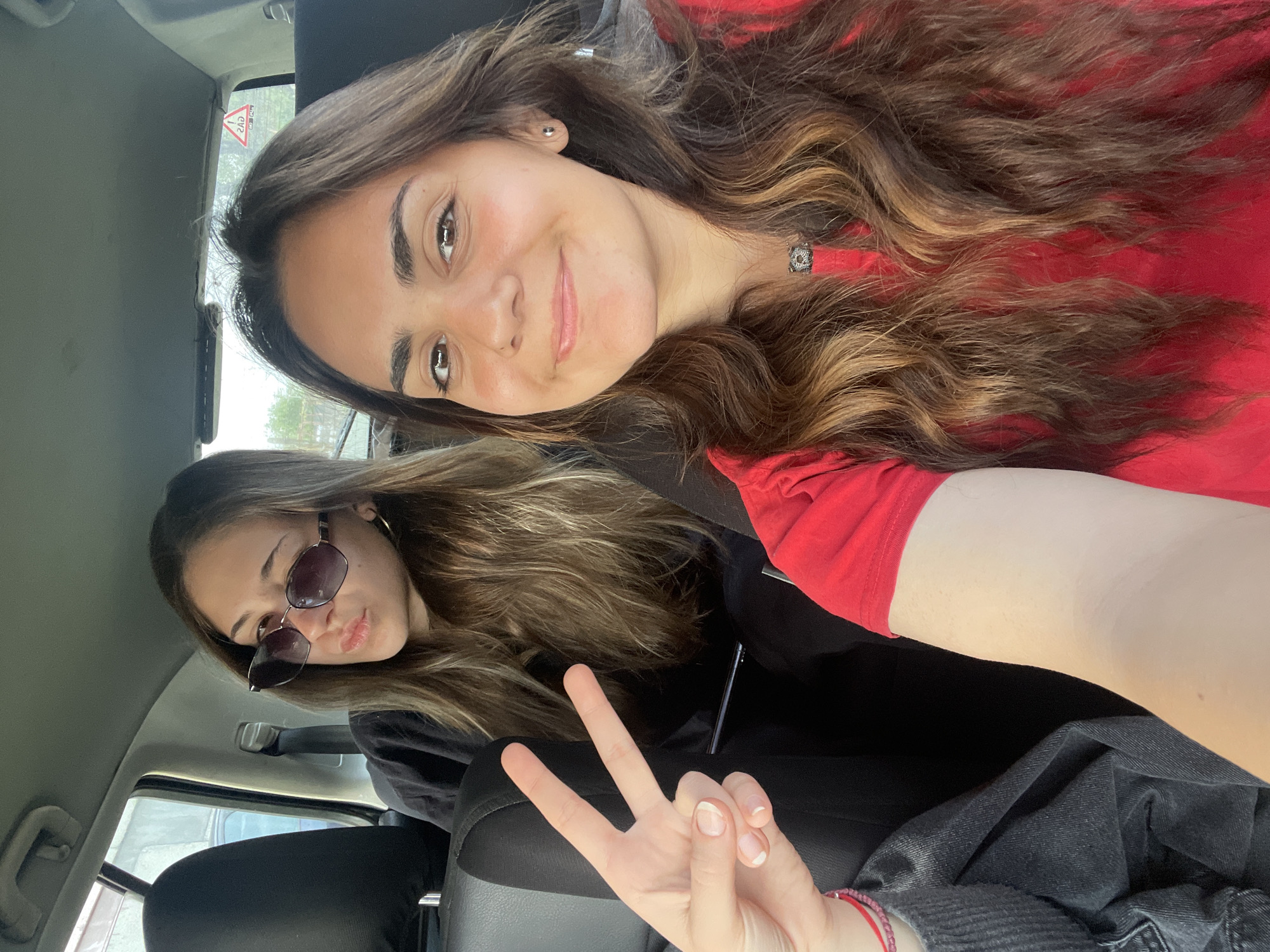Two young women are in a car, presumably taking a selfie. The woman in the foreground is wearing a red shirt and has long, brown hair with blonde highlights. She is smiling at the camera. The woman in the background is wearing sunglasses and a black shirt. Her hair is also long and brown with blonde highlights. A hand is making a peace sign in the bottom part of the image. The location is an interior of a car in the city of Unknown, Unknown.