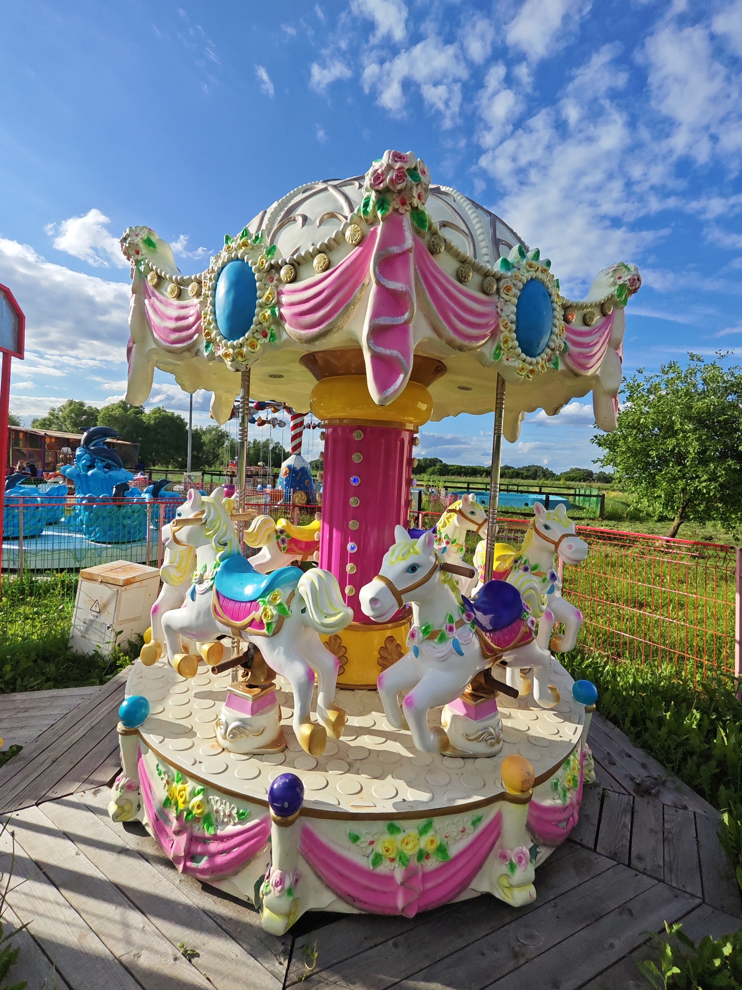This outdoor scene depicts a carousel located in Bronnitsy, Russia. The carousel features white horses with colorful saddles (blue, purple) and gold accents. The carousel's top is adorned with pink fabric, floral designs, and blue oval embellishments. The carousel rests on a wooden platform, surrounded by greenery. In the background, there is a dolphin-themed ride with blue water effects, as well as a red fence separating the carousel from an open field. The sky is blue with scattered clouds. The overall setting appears to be an amusement park or recreational area.