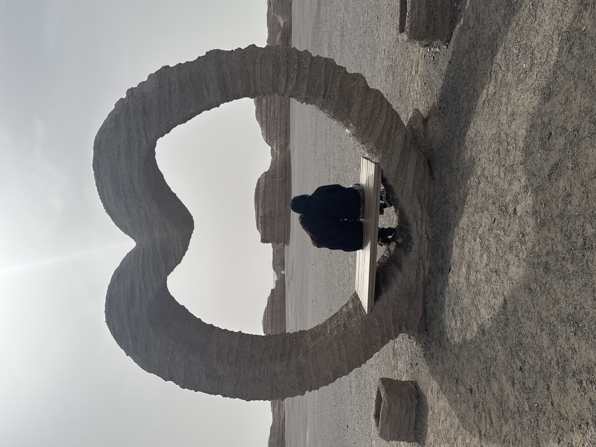 Two people are sitting on a bench within a stone heart-shaped structure. The scene is set within the Yardang National Geological Park, Dunhuang City, China. The landscape is arid and rocky. The sky is light.