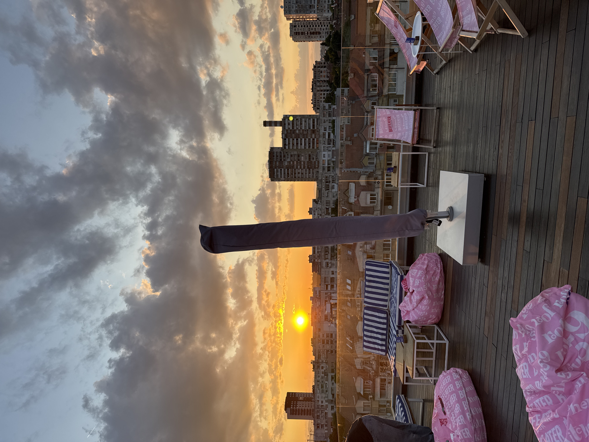 This image depicts a rooftop terrace in the city of Sofia, Bulgaria, at sunset. The sun, a bright orange disc, is prominently visible on the left horizon, casting warm, golden light across the scene. The sky transitions from a light blue at the zenith to vibrant orange and yellow near the horizon, populated by numerous dark grey and partially illuminated clouds. A faint aircraft contrail is visible in the upper-left sky.
The rooftop features dark wooden deck flooring. In the foreground and mid-ground, various seating arrangements are present. These include two-person sofas with blue and white striped cushions and white metal frames, accompanied by small, square, white-framed tables with light wooden tops. Several large, light pink beanbags, emblazoned with white "BEEFEATER LONDON" and "BEEFEATER PINK" text, are scattered across the deck. To the right, multiple light pink fabric deck chairs with wooden frames, also branded "BEEFEATER LONDON," are arranged around small, round white tables. A tall, grey, fabric-covered object, likely a folded large umbrella or canopy, stands on a white square base in the center-right. A clear glass railing defines the edge of the terrace, reflecting the sunset and the urban landscape.
The urban environment of Sofia stretches into the background. Immediately below the terrace, residential buildings with reddish-brown tiled roofs are visible. Further in the distance, a dense array of multi-story buildings, including prominent grey communist-era panel blocks, forms the city skyline. A tall, dark, multi-story building is silhouetted on the left horizon, and a large complex of buildings with a distinct, slender, dark industrial chimney is visible in the mid-right. One person is partially visible on the far left, seen from behind, wearing blue jeans and a dark top.