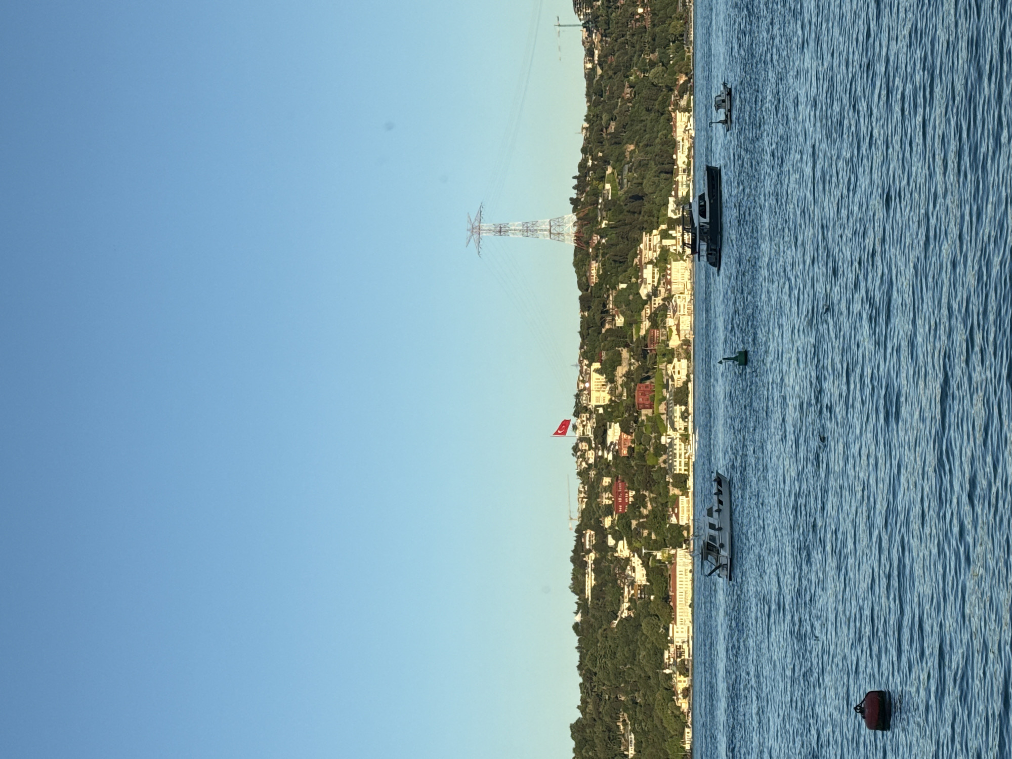 The image shows a scene on the Bosphorus strait in Beşiktaş, Turkey. The right side of the image is the water, which is rippled, with several floating objects: a red buoy in the lower right corner, a green navigational buoy near the shoreline, and several boats of varying sizes. The left side of the image is the coastline, with buildings scattered amongst trees. A Turkish flag is seen flying on the hill. An electrical transmission tower is visible in the distance. The sky is clear blue.