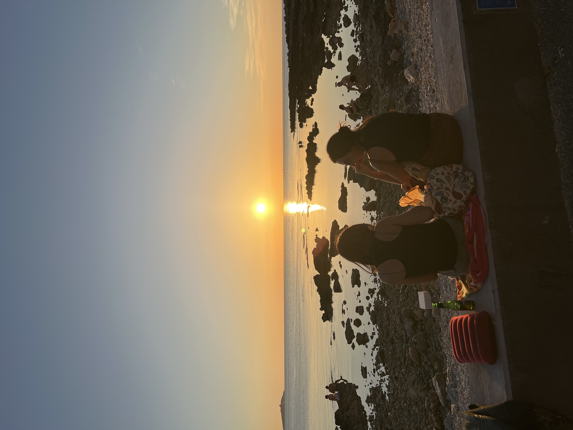 Two individuals are seated on a concrete ledge facing a body of water at sunset. The sky is a gradient of pale blue and yellow, with the sun setting on the horizon, casting a golden reflection on the water. The foreground shows a rocky shoreline with some of the two individuals' belongings, including a red cushion and a bottle of beer. In the background, there are other people visible on the rocks and in the water. The overall scene depicts a tranquil evening by the sea.
