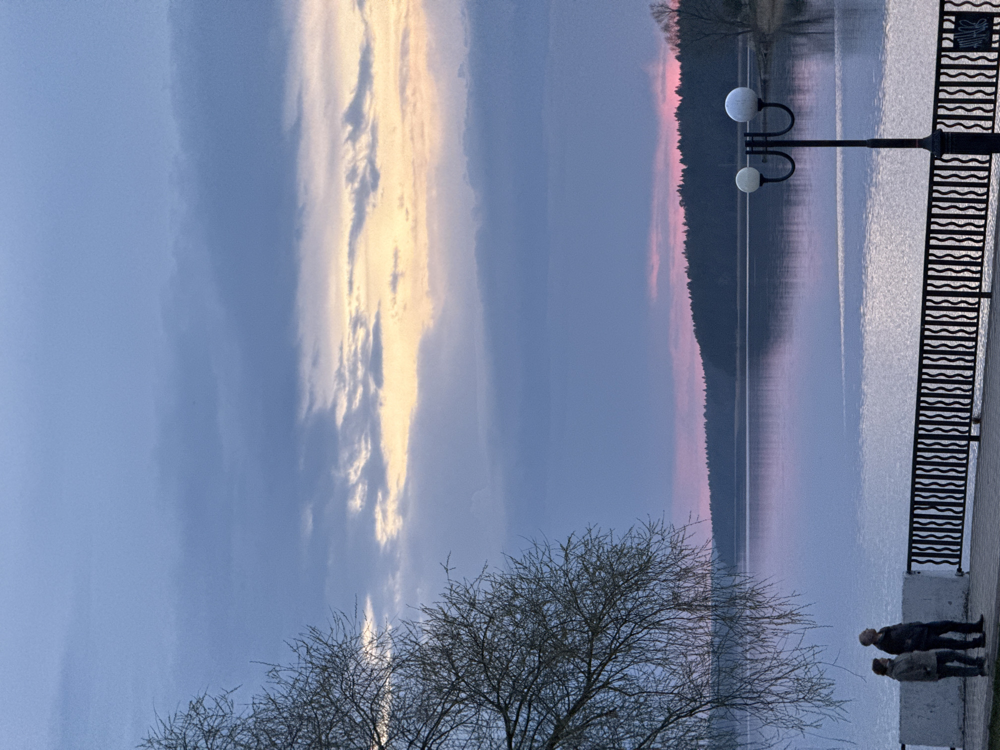 Captured in Votkinsk, Russia, the image presents a landscape scene at dusk or dawn, with the sky painted in shades of blue, pink, and gold. Wispy clouds stretch across the upper portion of the frame. The scene includes a body of water that mirrors the colors of the sky, creating a serene reflection. A dark treeline runs along the horizon, providing a stark contrast to the pastel shades of the sky. On the right side, a portion of a city embankment is visible, featuring a wrought-iron railing and a lamppost with two globe-shaped lights. At the lower right, two figures stand near the edge of the embankment, appearing to observe the view. In the foreground, bare tree branches reach upwards, silhouetted against the sky.