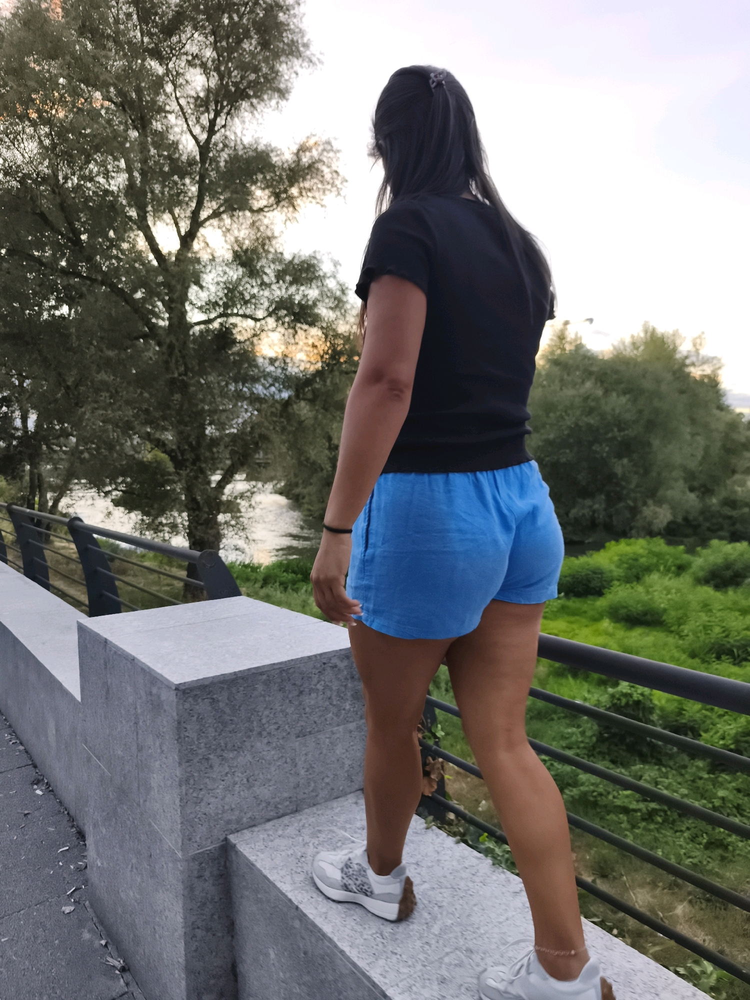 An unidentified woman, viewed from behind and to her left, walks along a concrete railing outdoors. She has long, dark hair and wears a black t-shirt and bright blue shorts. Her right arm is bent at the elbow, with a black bracelet on her wrist. She wears white athletic shoes. The background features a body of water, trees, and green foliage. The scene appears to be a park or a waterfront area.