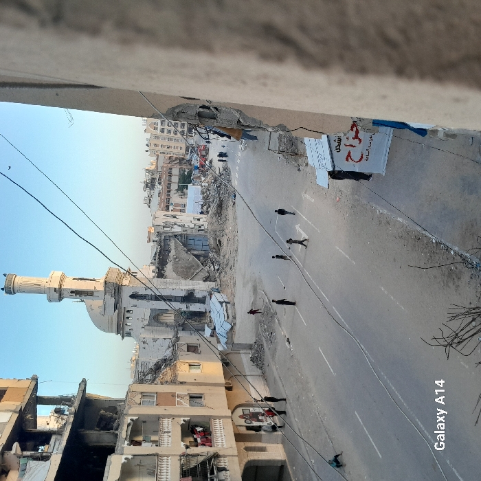The image displays a damaged urban streetscape in Khan Yunis, Palestinian Territory, observed from an elevated vantage point, likely during morning or late afternoon. A prominent mosque with a minaret and dome, exhibiting structural damage and surrounded by extensive rubble, anchors the background. To the left, a multi-story residential building displays significant facade damage with exposed interiors; several individuals are gathered at its ground level and on balconies, with laundry visible on some balconies. A paved street with two marked lanes runs through the foreground and midground, flanked by dirt and debris. Approximately 15 individuals are present: a cluster near the damaged building on the left, several males distributed across the street (some appearing to be playing), and others further down the thoroughfare, some near a white tent structure. Overhead utility wires crisscross the upper frame. On the right, a partial building structure bears a white banner with Arabic text, including "تسجيل" (registration) and "جراح" (wounds/surgery). The sky is clear.