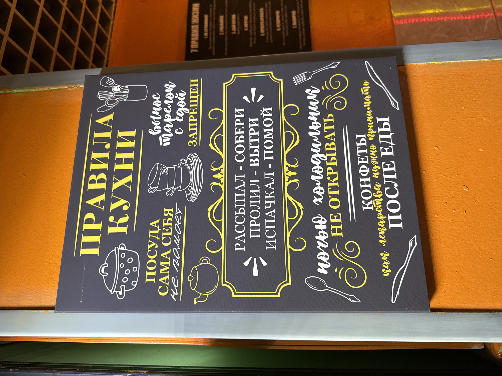 The image shows a close-up of a dark grey sign with yellow lettering and white line drawings of kitchen items. The sign has several rules written on it in Russian. The most prominent text at the top reads "ПРАВИЛА КУХНИ" (Rules of the Kitchen). Below this, there are several points with illustrations: a pot, a stack of plates, a teapot, and a holder with kitchen utensils.
The rules on the sign translate to:
- "ПОСУДА САМА СЕБЯ не помоет" (Dishes won't wash themselves)
- "вынос тарелок с едой ЗАПРЕЩЕН" (Taking plates with food out is forbidden)
- "РАССЫПАЛ - СОБЕРИ. ПРОЛИЛ - ВЫТРИ. ИСПАЧКАЛ - ПОМОЙ" (Spilled - collect. Spilled liquid - wipe. Soiled - wash)
- "НОЧЬЮ ХОЛОДИЛЬНИК НЕ ОТКРЫВАТЬ" (Do not open the refrigerator at night)
- "КОНФЕТЫ как лекарства нужно принимать ПОСЛЕ ЕДЫ" (Sweets, like medicine, should be taken after meals)
To the right of this sign, a smaller, darker sign with red and white text is visible, which appears to list "7 ПРАВИЛ ЖИЗНИ" (7 Rules of Life). The background of the main sign is set against an orange wall.
Based on the presence of the sign and the style of the decor, this scene could be from a cafe, a restaurant, or a communal kitchen area. Without further contextual information or other identifying landmarks, it is impossible to pinpoint the exact location within Saint Petersburg, Russia. However, the style of the signage and the language suggest a location within Russia.