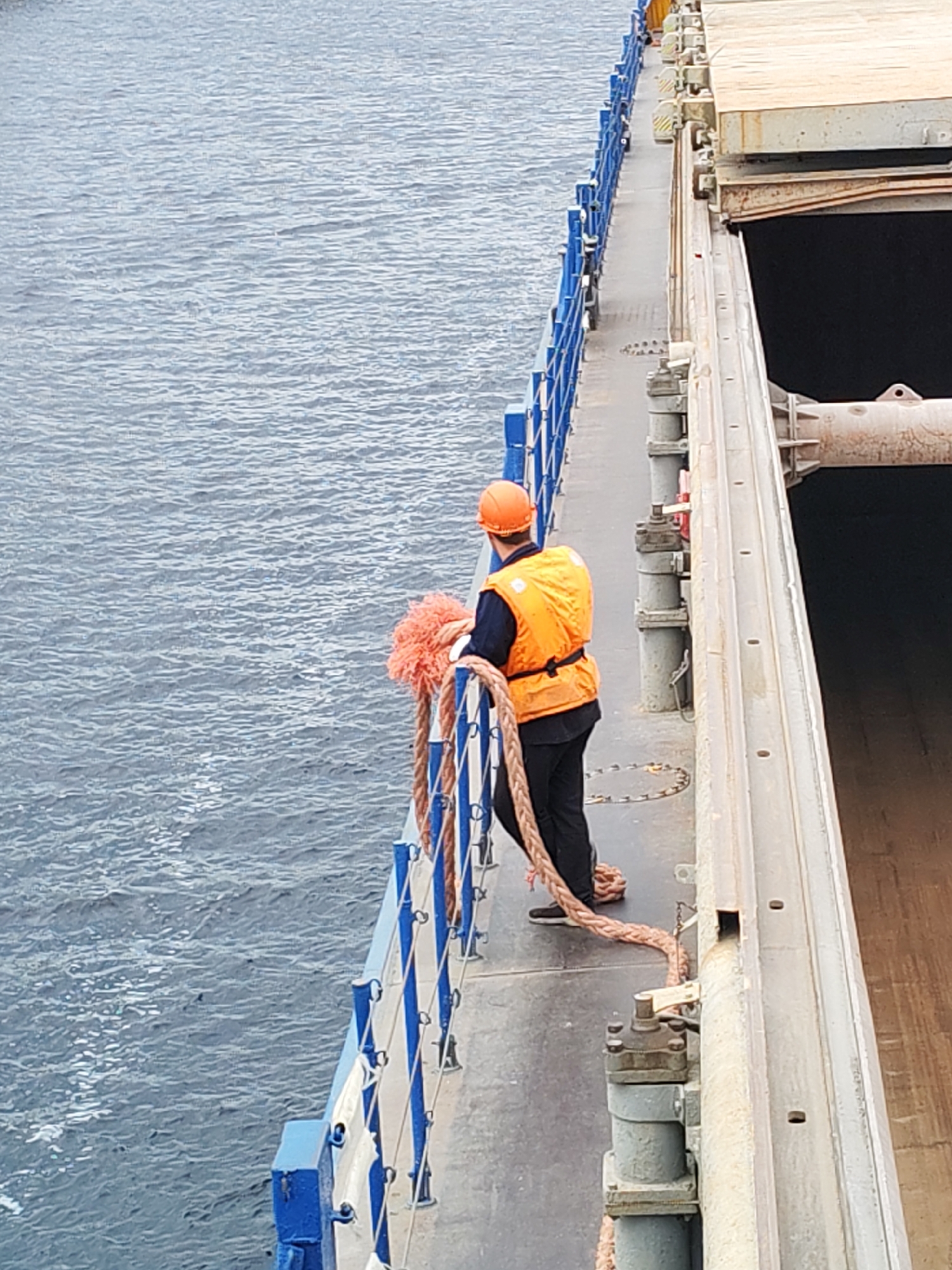 A person wearing an orange hard hat and a bright orange life vest stands on the deck of a ship, holding a thick rope. The person is wearing dark pants and dark shoes. The ship's deck has a blue railing along the edge. To the right of the deck is a large, open cargo hold with wooden planks. The sea, with visible ripples, surrounds the ship. The image appears to be taken from a higher vantage point, looking down on the scene. The location is identified as Shal'skii, Russia.